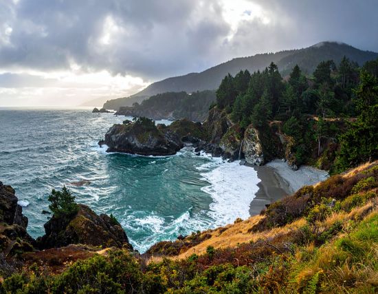 Rocky ocean cove with crashing waves, forested cliffs, and misty hills under a cloudy sky.