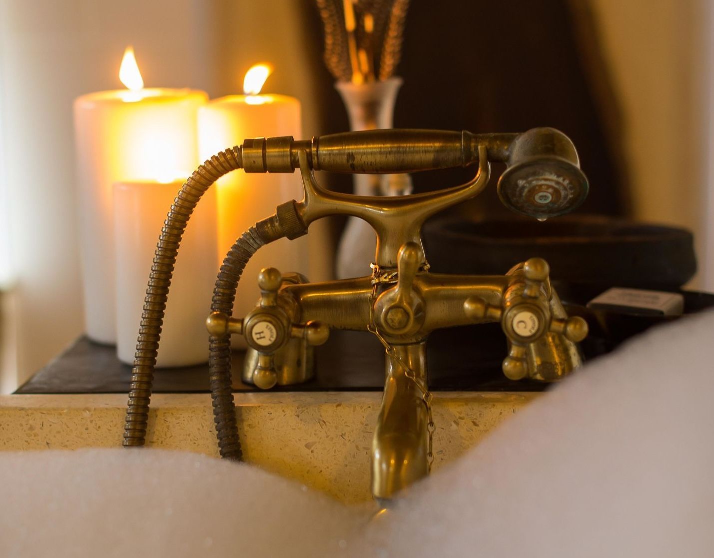 Vintage brass bathtub faucet with coiled hose beside lit candles on a bath shelf