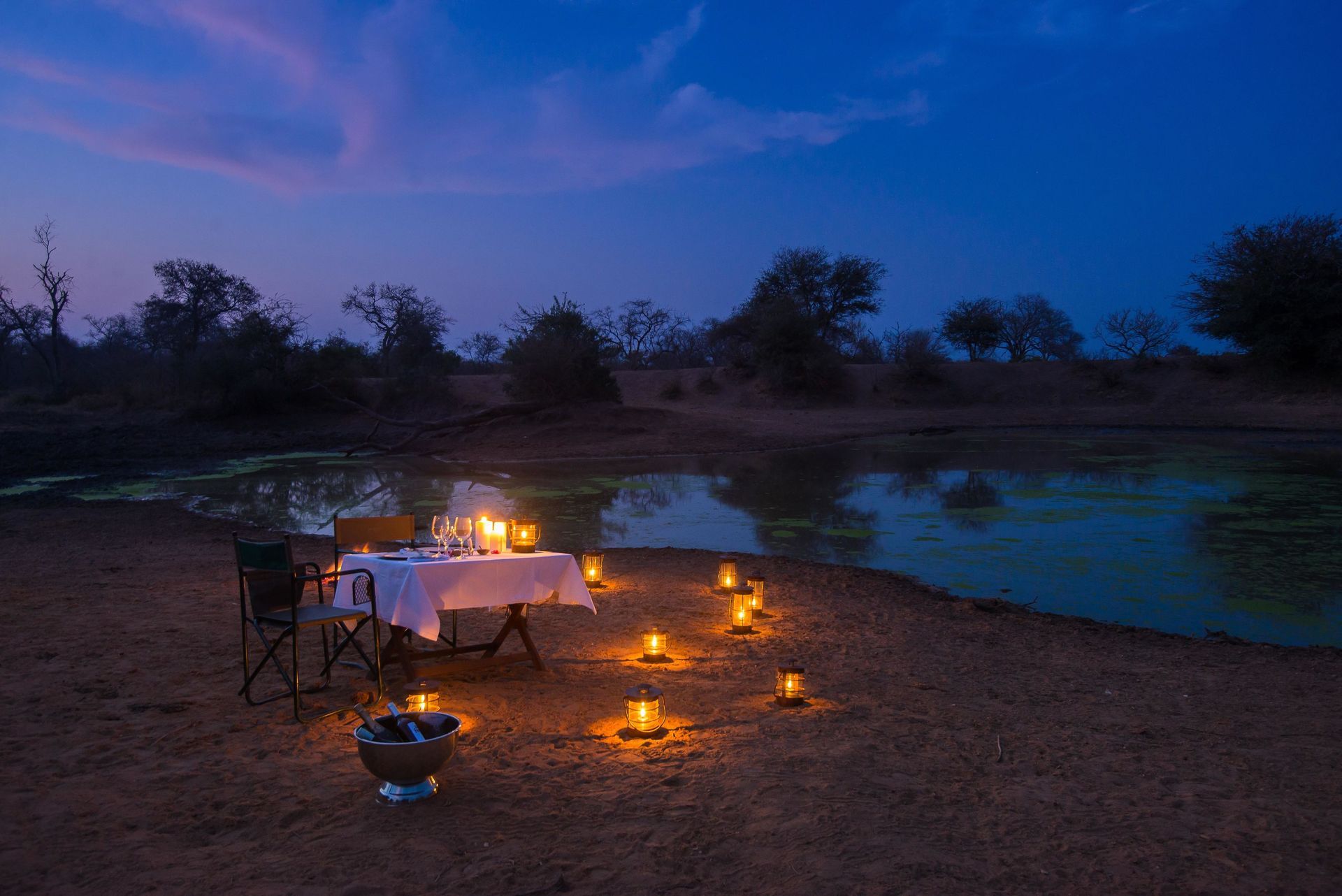 Lit dinner table and campfire by a lake at dusk, with candles glowing under a blue sky.