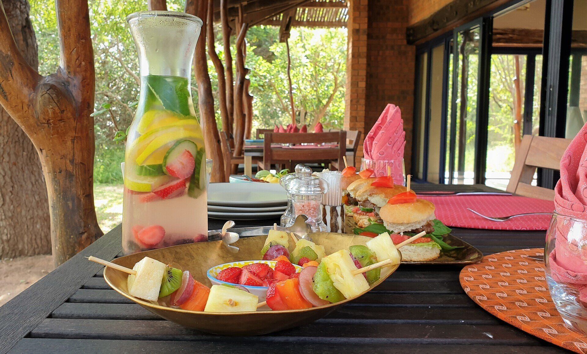 Fruit and dessert plates on a wooden table in a rustic outdoor dining area with a pitcher of drink