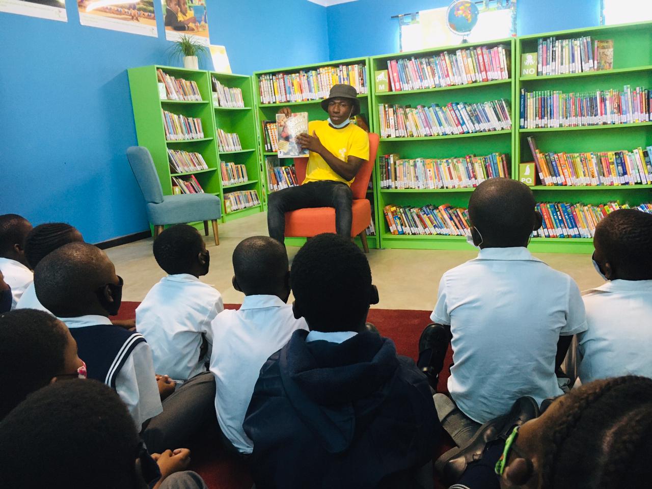 Children seated on the floor listen to a speaker in a colorful library with green bookshelves.