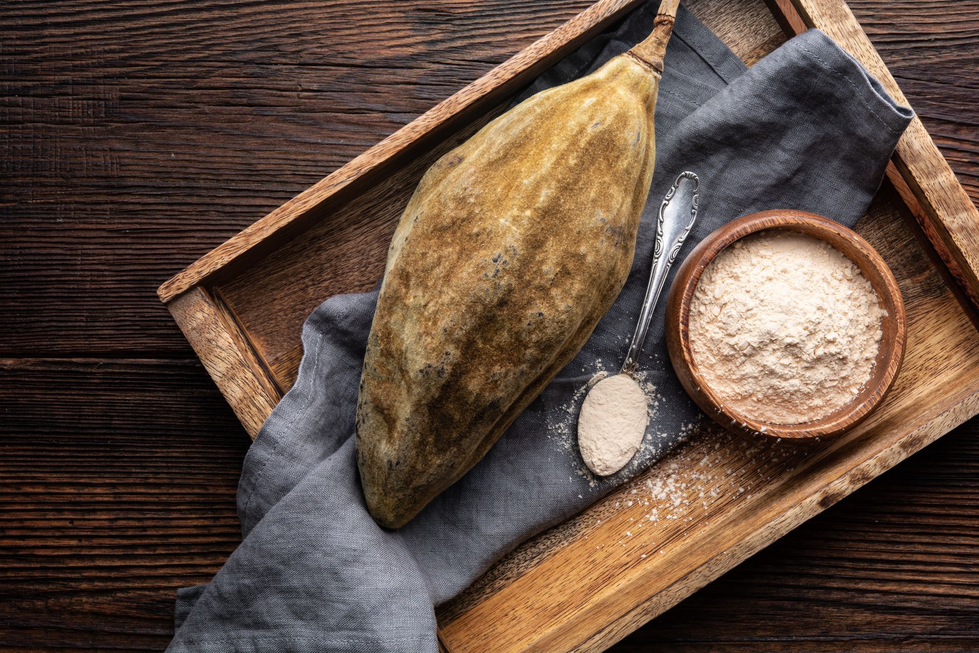 Cacao pod, powder, and spoon on a wooden tray with a gray cloth on a rustic table