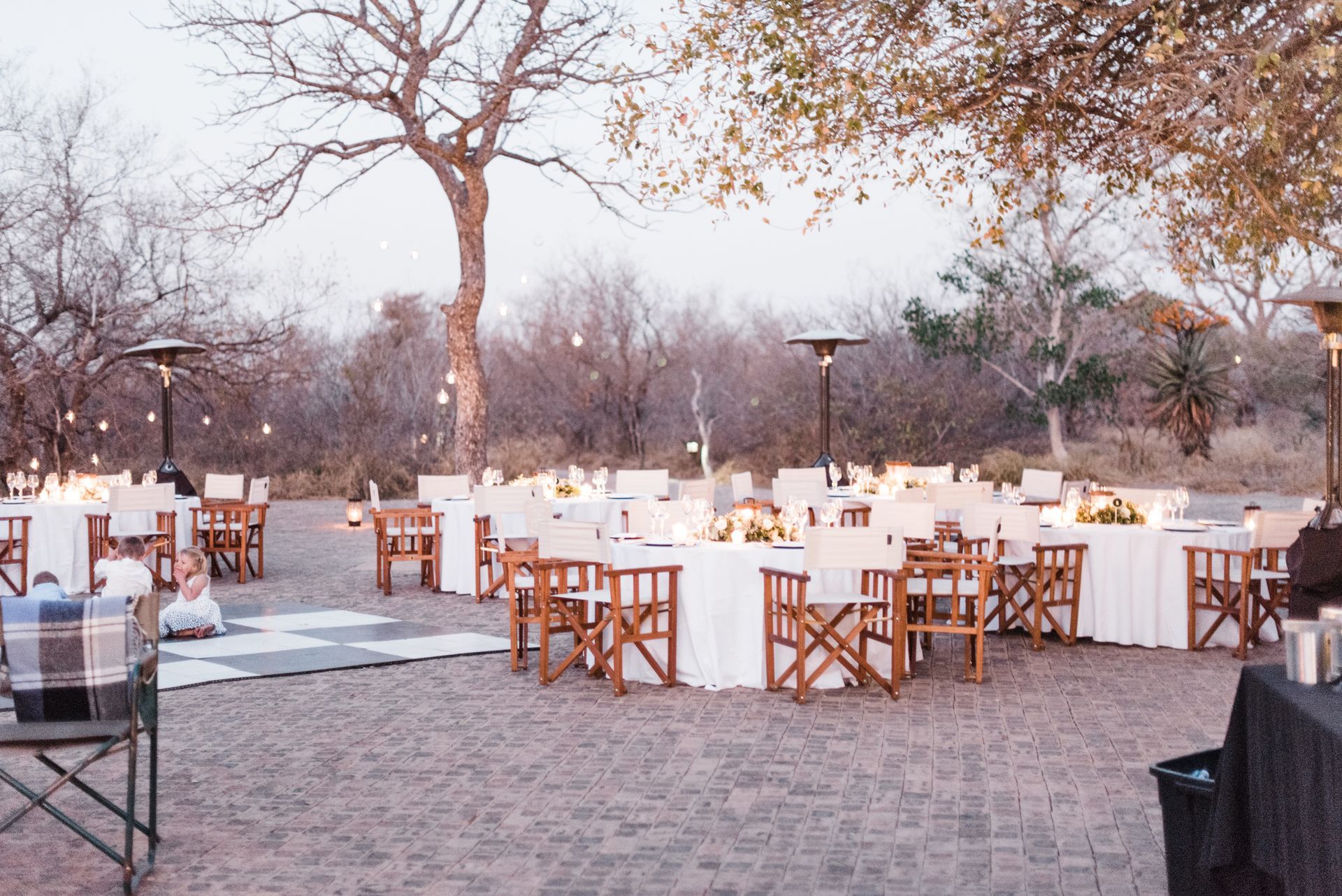 Outdoor dining setup with white tablecloths and wooden chairs on a sandy terrace under trees