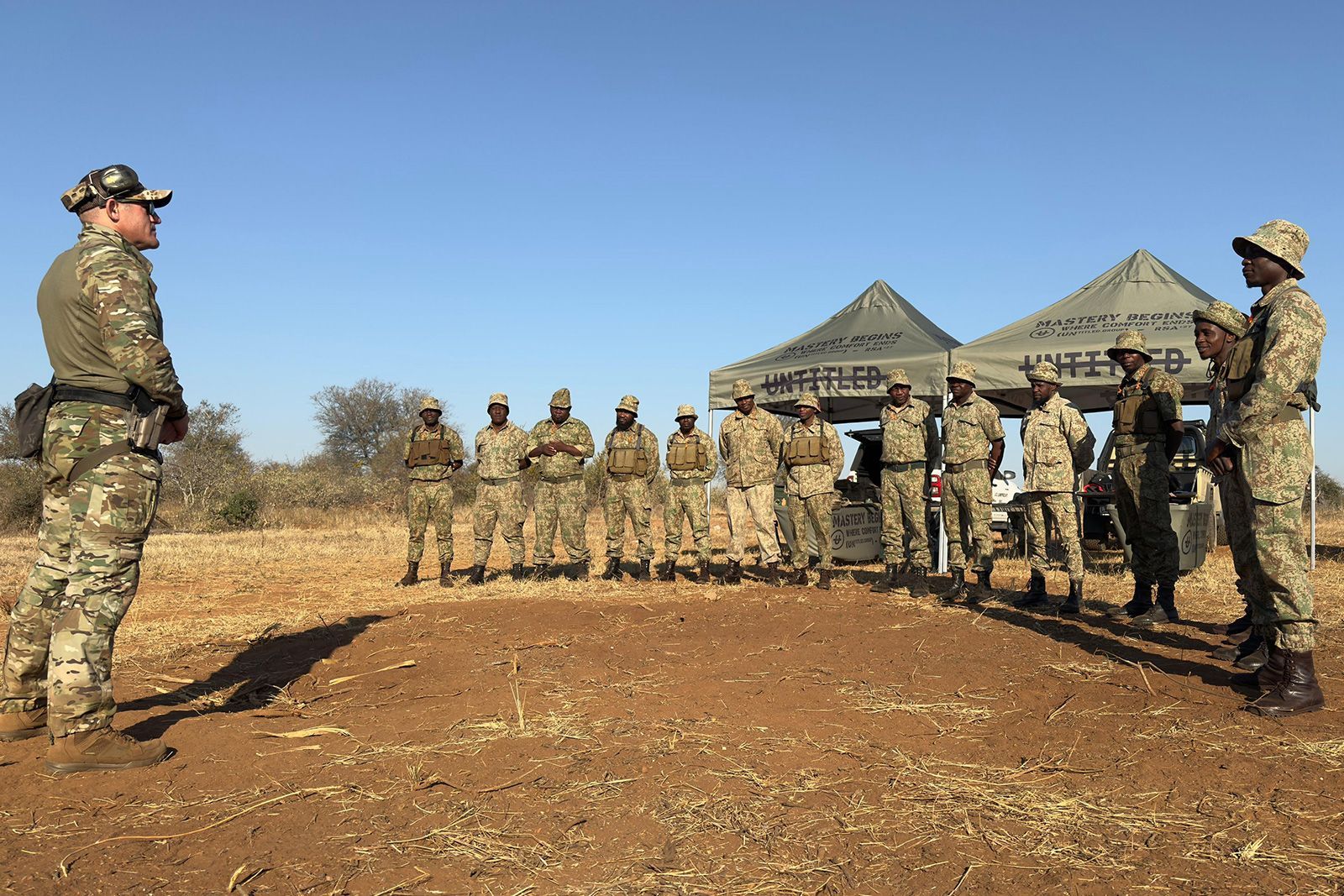 Soldiers standing in formation outdoors near tents in a dry, open field under a clear blue sky