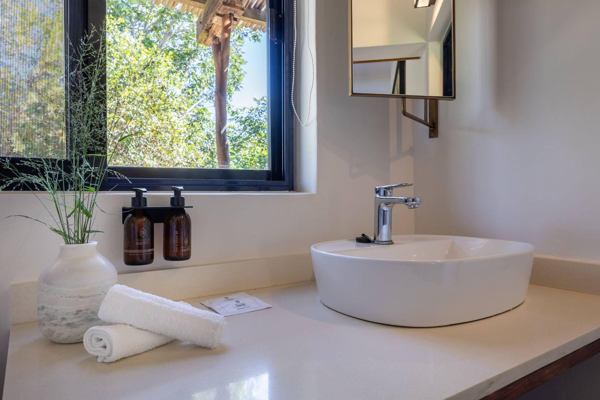 Minimalist bathroom vanity with white sink, mirror, and window view, with rolled towels and soap dispensers