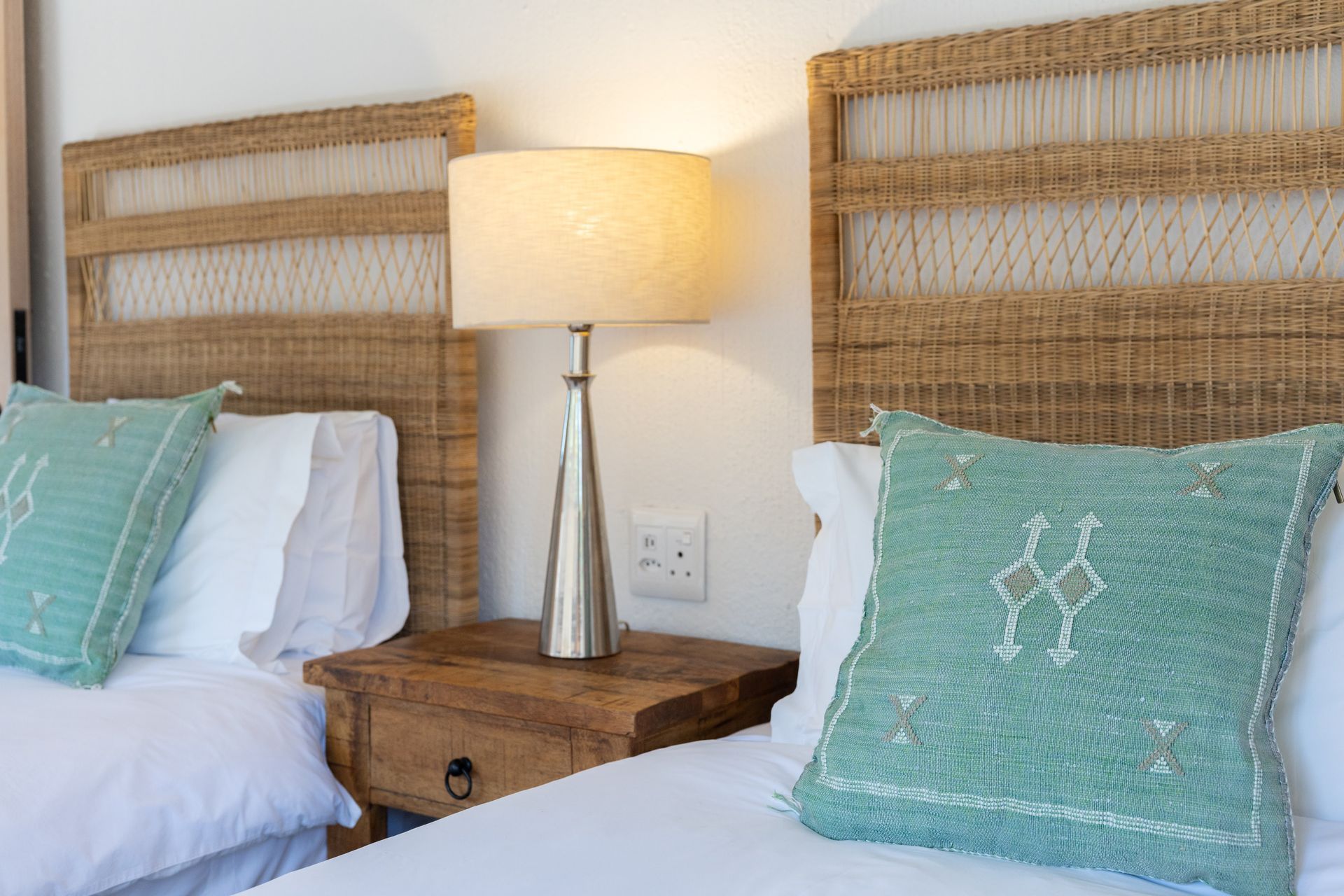 Two twin beds with teal pillows, a bedside lamp, and a wooden nightstand in a bright bedroom.