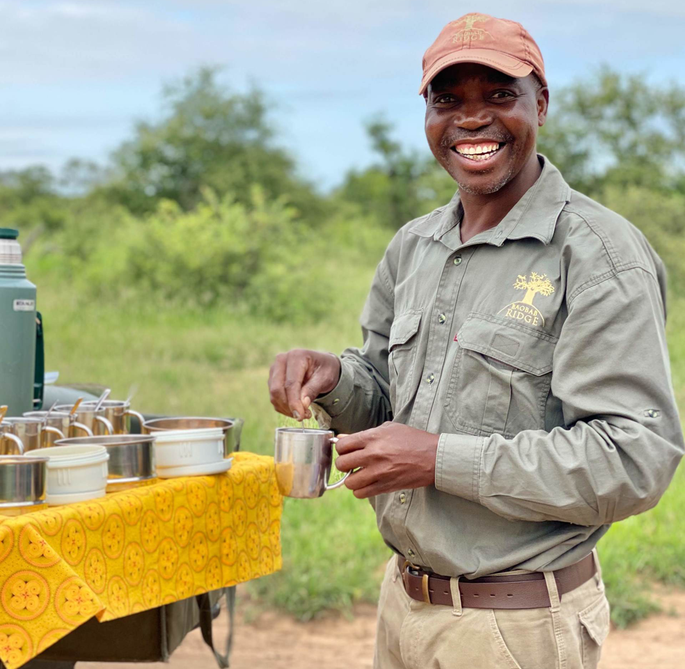 Smiling person serving tea outdoors beside a table with metal cups and a yellow cloth