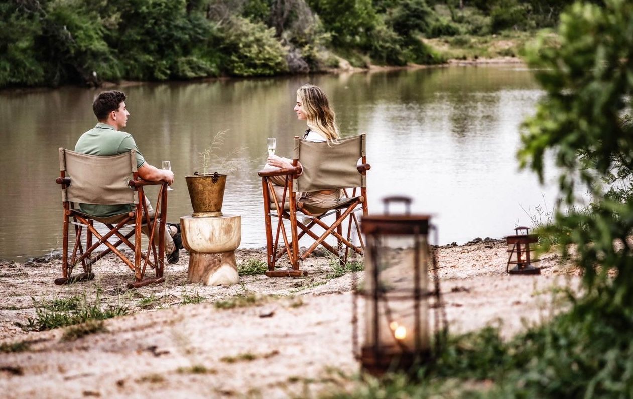 Two people sitting in folding chairs by a river, with lanterns and a small table in a wooded setting