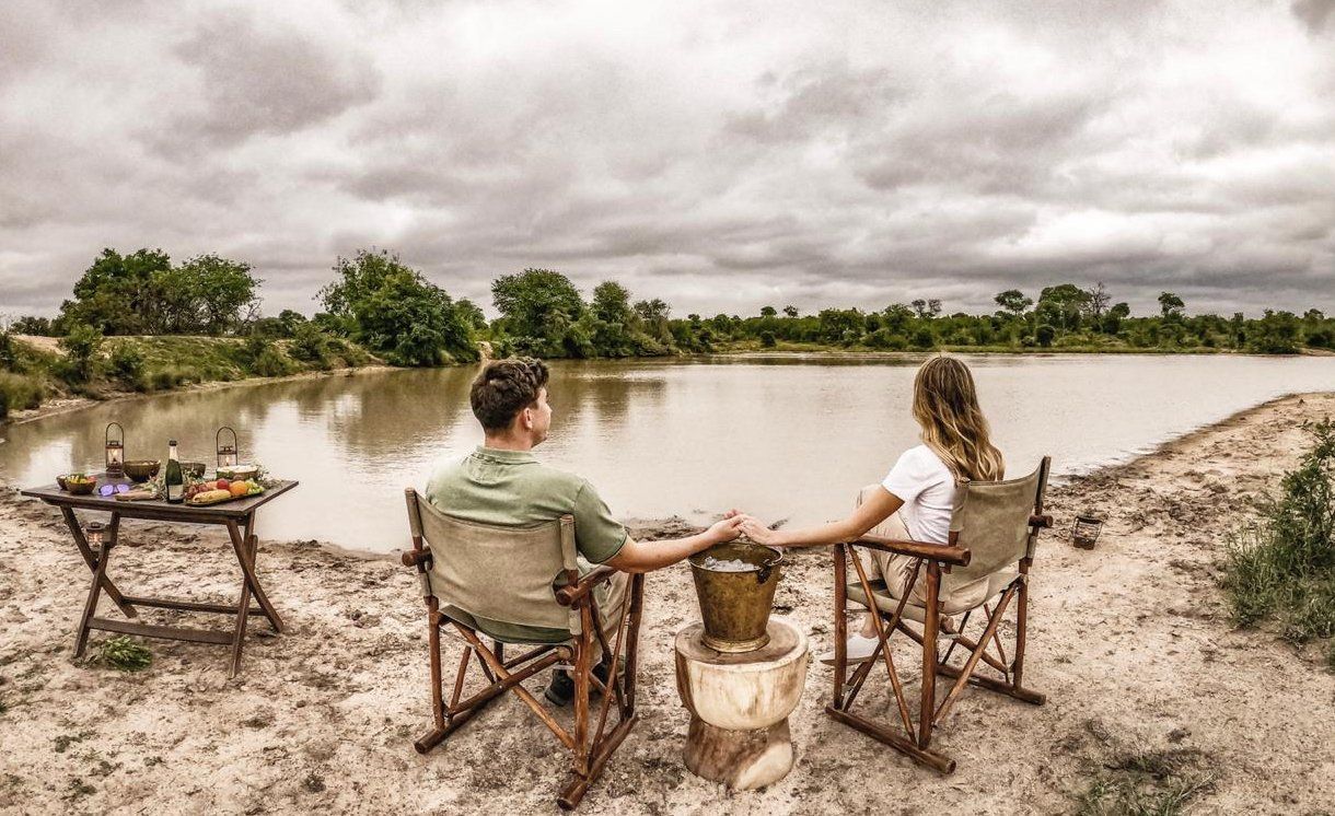 Two people sit by a muddy river, facing a cloudy sky over a grassy wetland.