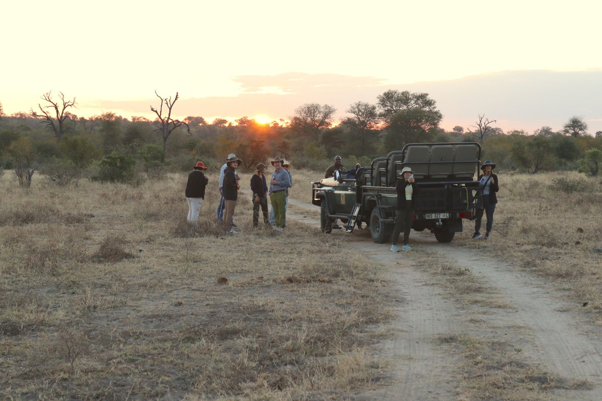 People gathered near safari vehicles on a dirt road at sunset in a dry savanna.