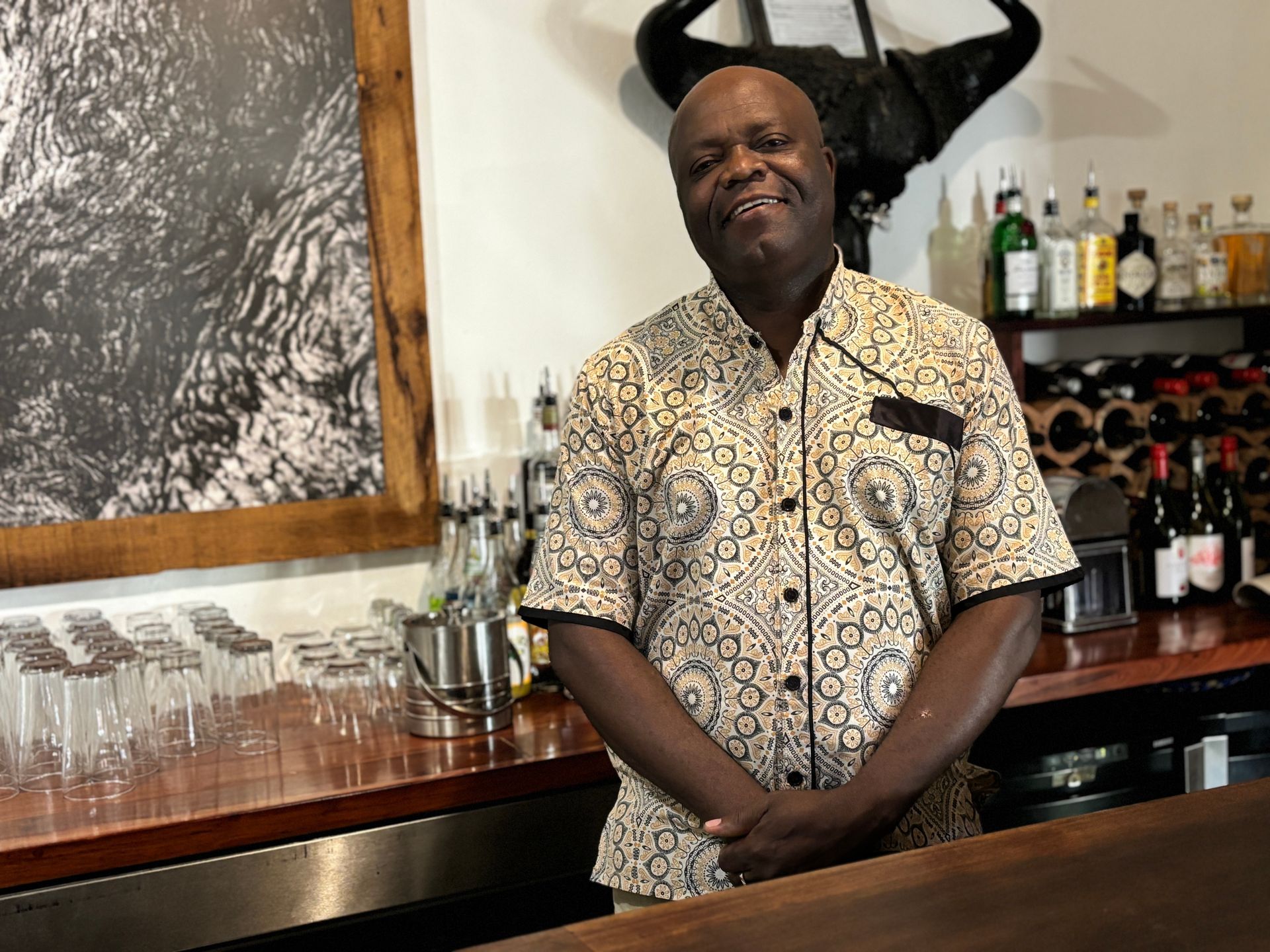 Man standing behind a bar, wearing a patterned shirt, with glasses and bottles on the shelves behind him.
