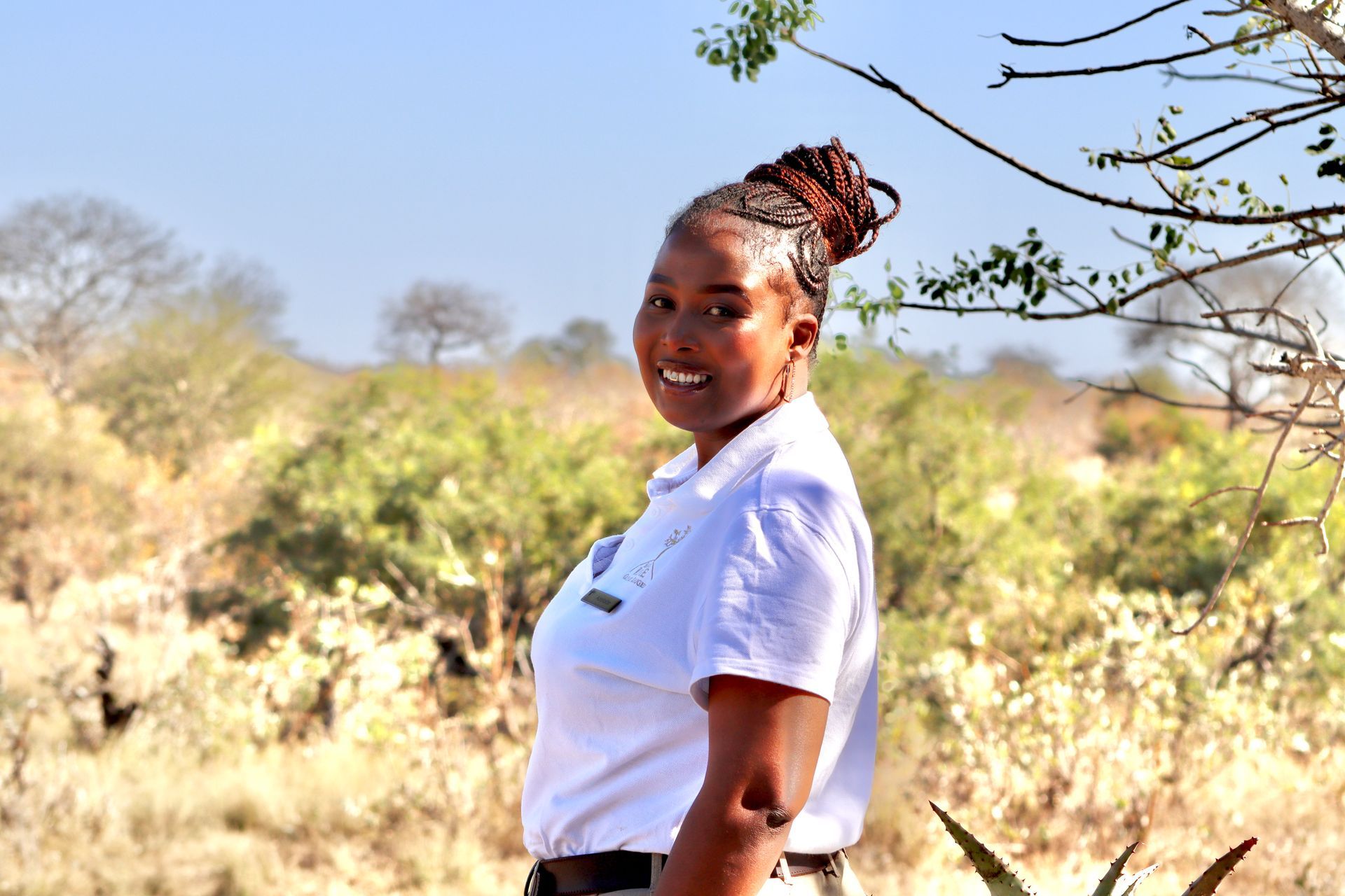 Person smiling outdoors in a white shirt, standing in a sunny grassy landscape.