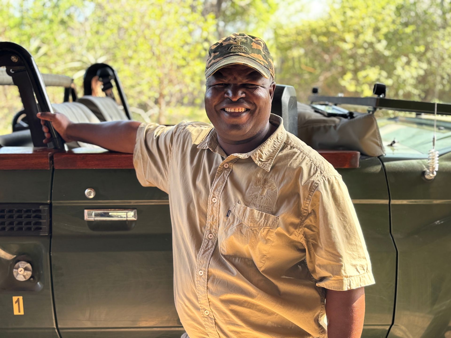 Smiling man in beige shirt and cap leaning on green safari vehicle outdoors