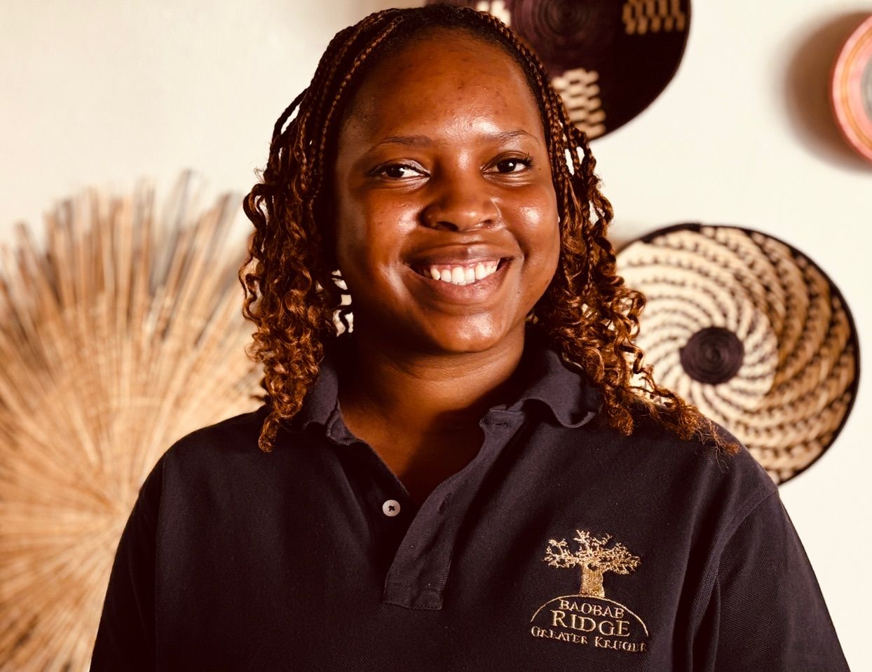 Smiling person in a black polo shirt standing indoors with woven wall decor in the background