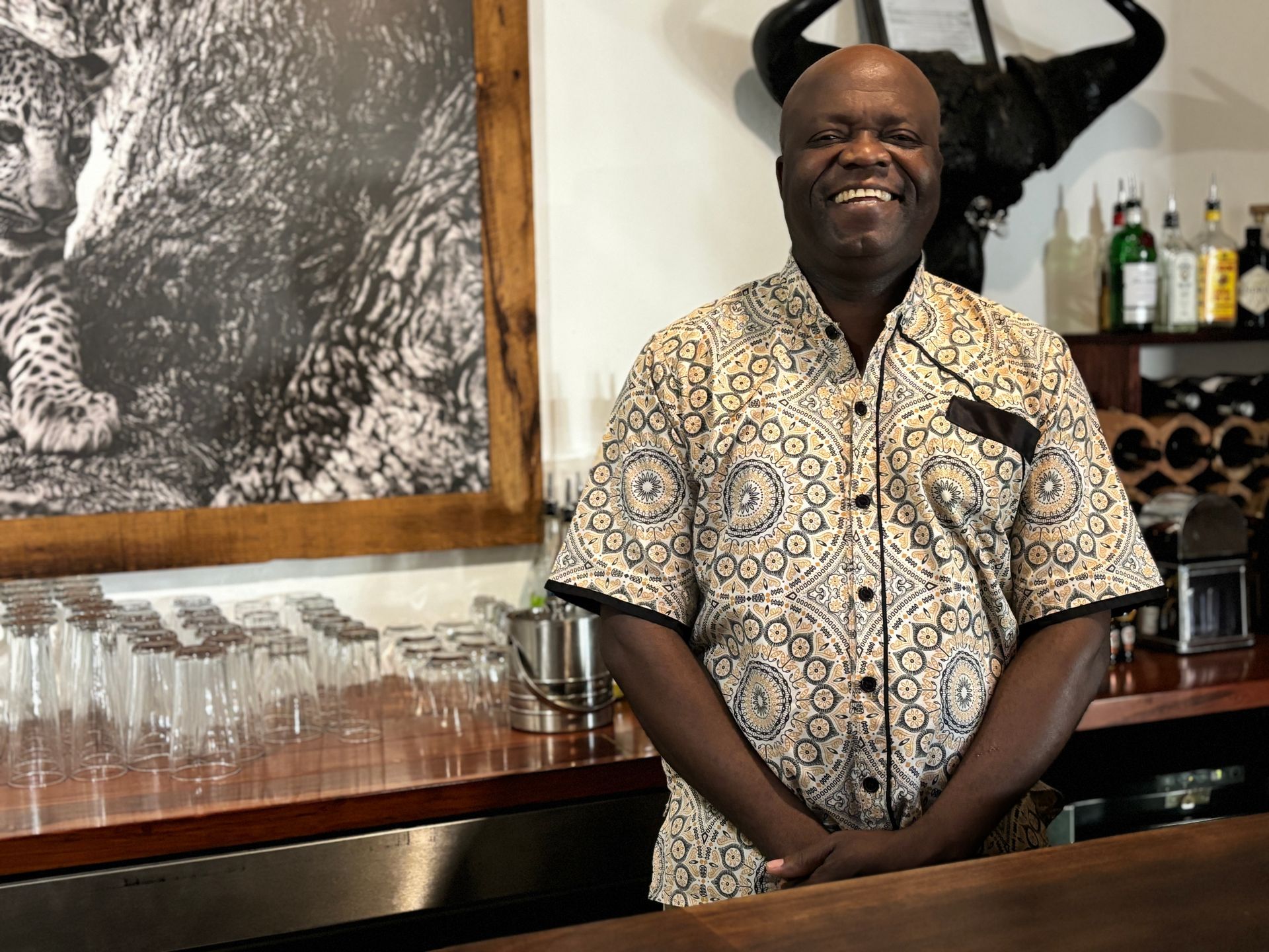 Man standing behind a bar counter, smiling, with hanging glasses and bottles on shelves behind him.