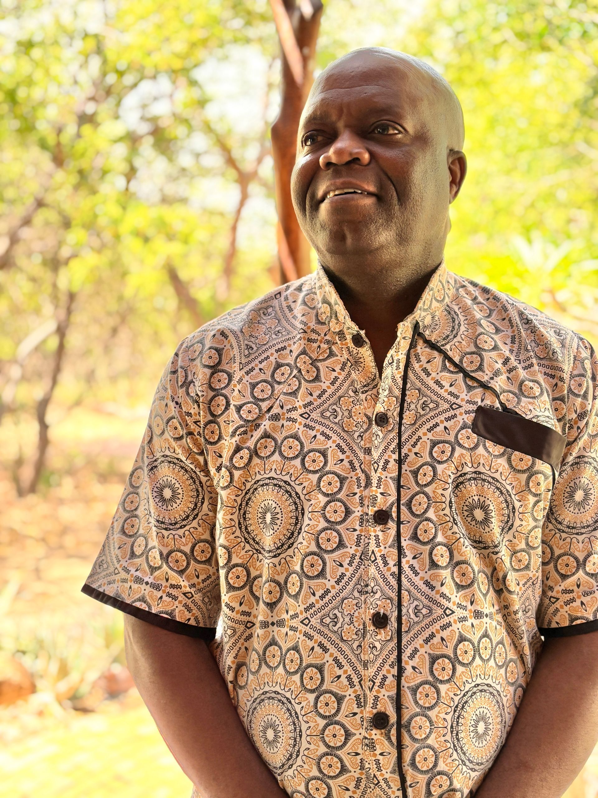 Man smiling in a patterned shirt standing outdoors with trees in the background
