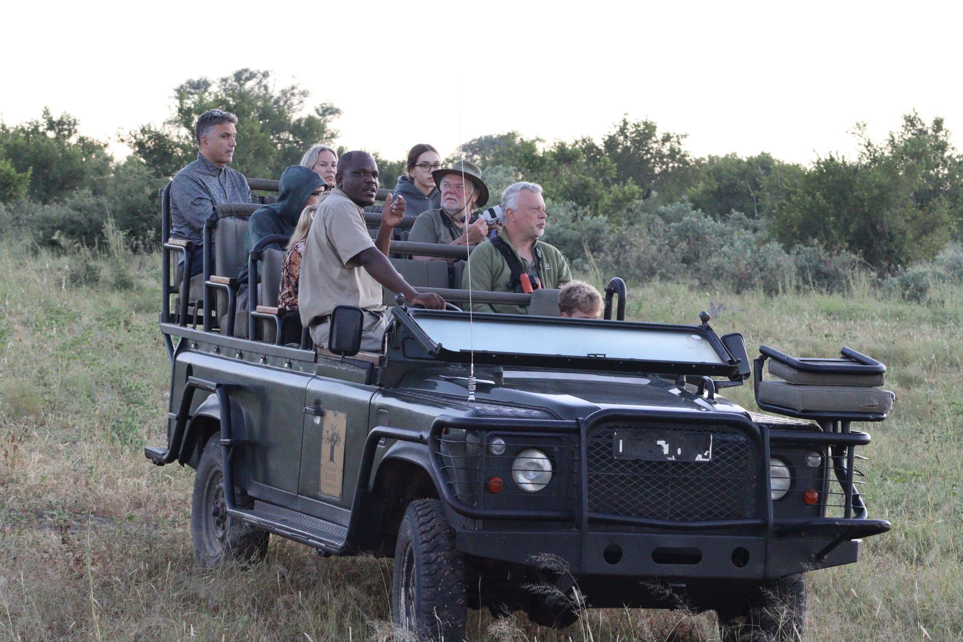 Open-top safari vehicle with several passengers driving through grassy savanna at dusk