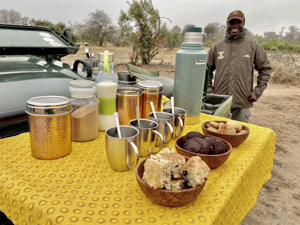 Outdoor table with jars, mugs, and bowls of food beside a thermos and a person in a cap at camp.