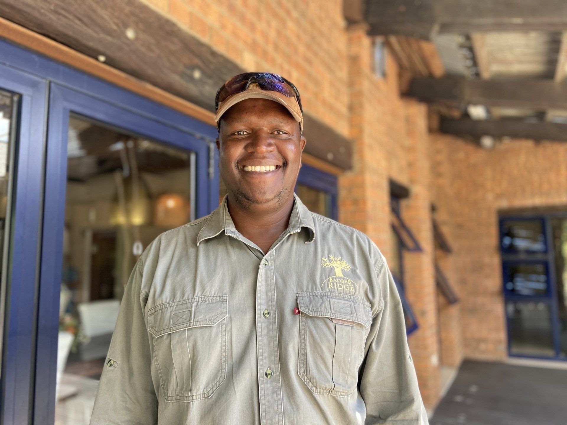 Smiling person in a cap stands outside a brick building with blue-framed windows.