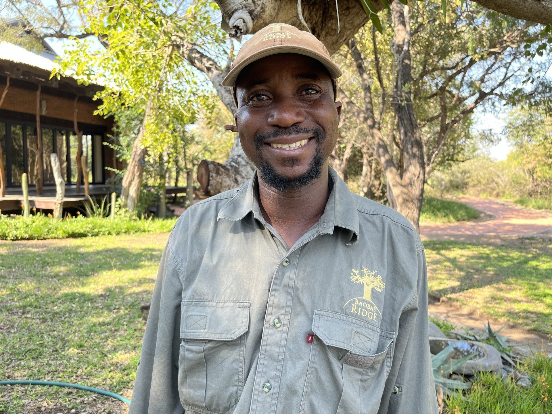 Smiling man in a tan cap and gray shirt standing outdoors in a sunny garden.