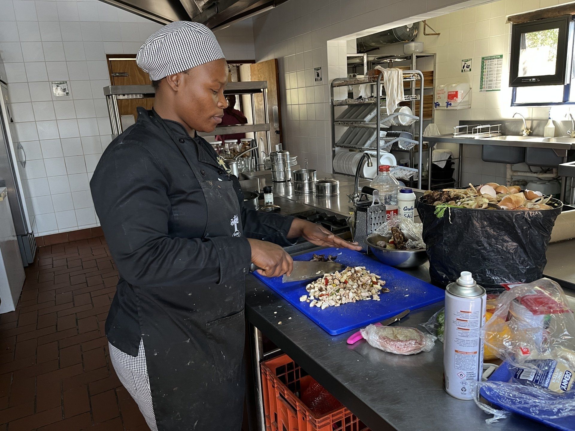 Chef cooking at a stainless-steel counter in a commercial kitchen, chopping food on a blue cutting board.