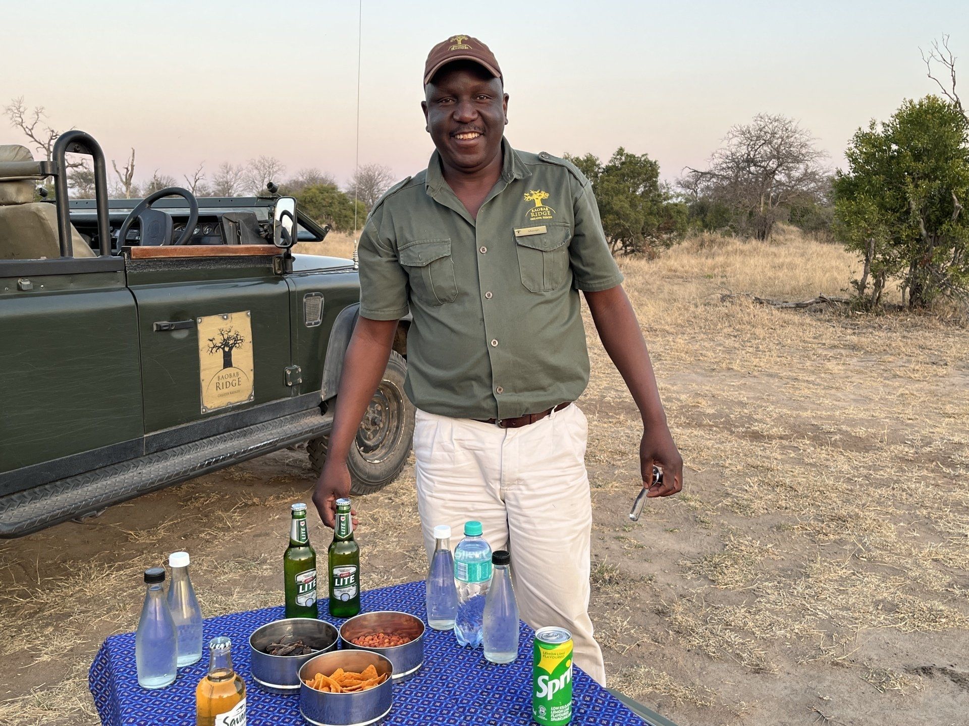 Man standing beside a table with bottles and bowls outdoors near a safari vehicle in dry grassland.