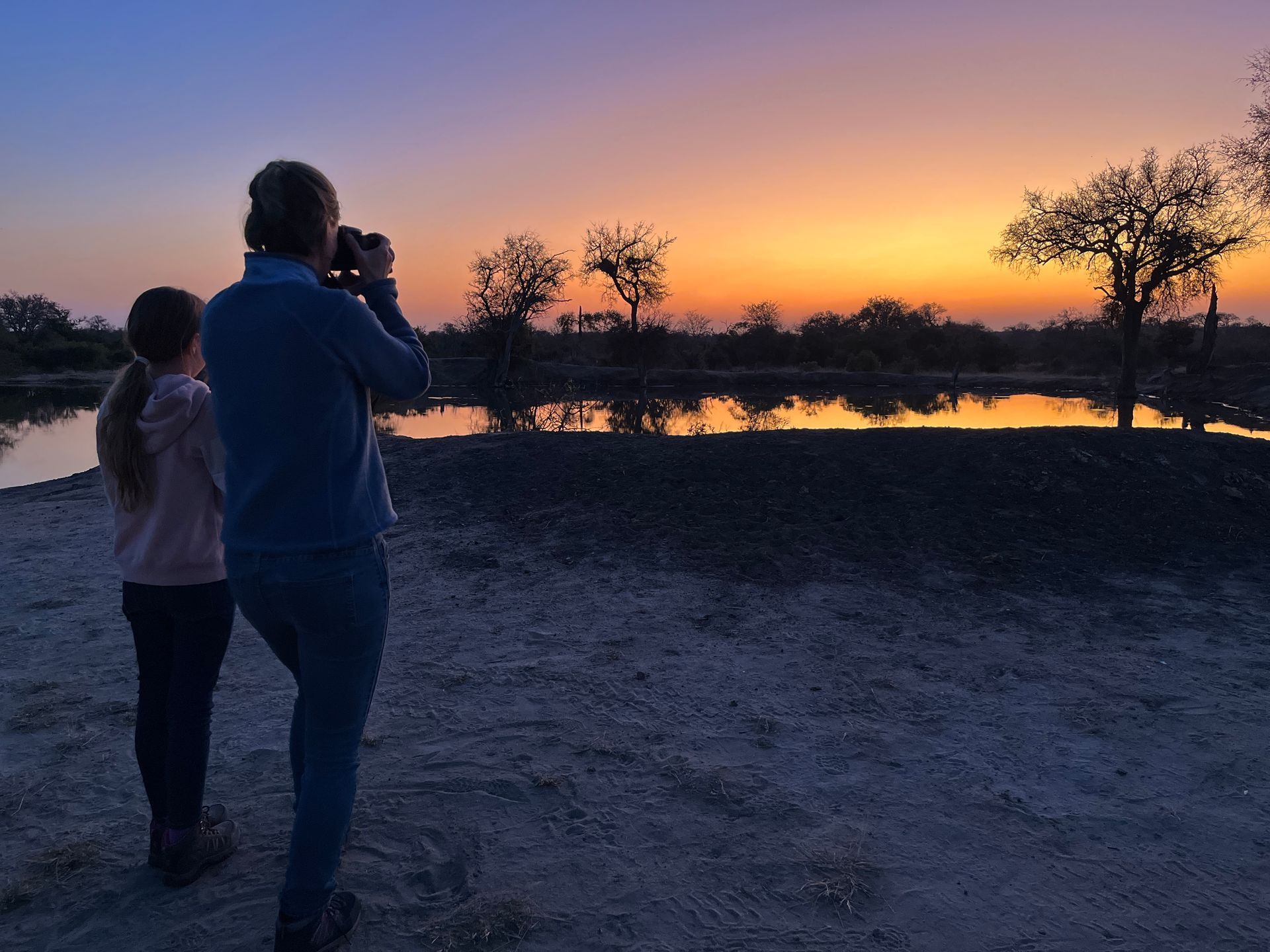 Two people photographing a sunset over water with silhouetted trees in the background.