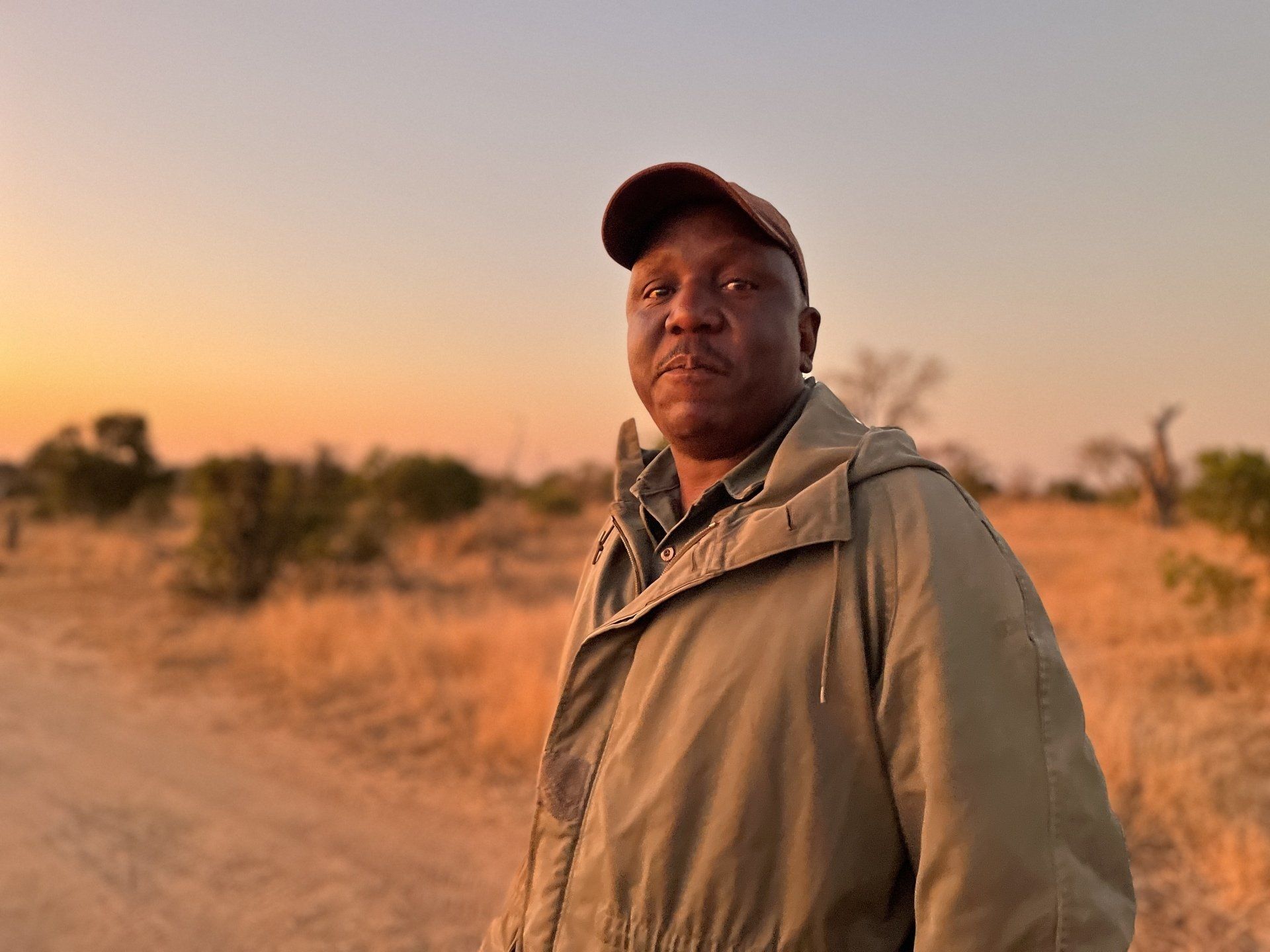 Person in a cap and tan jacket standing on a dirt road at sunset.