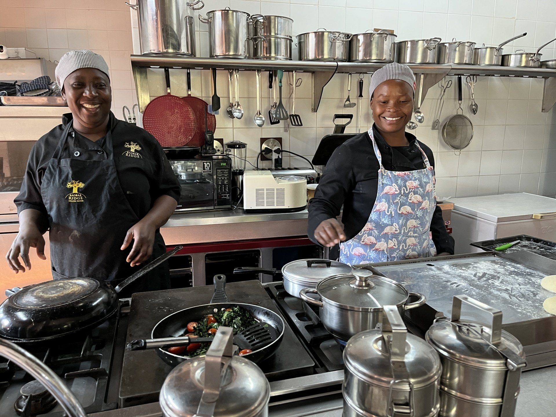 Two chefs smiling in a stainless-steel kitchen, cooking at a counter with pots, pans, and trays.