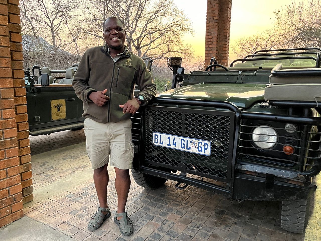 Man standing beside a black off-road vehicle in a driveway, wearing a green jacket and khaki shorts.