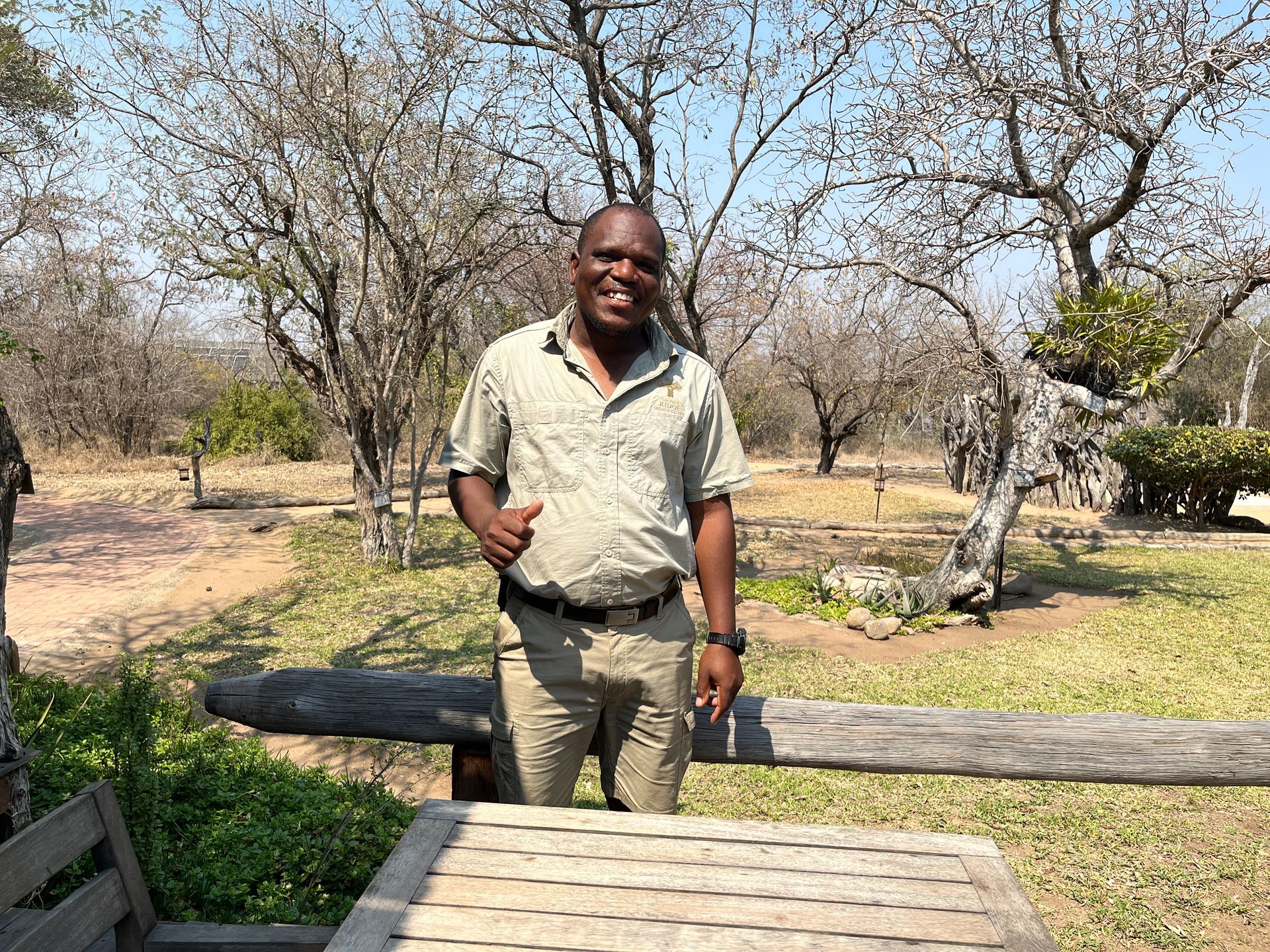 Man standing outdoors beside a wooden bench in a dry, tree-filled park, smiling at the camera