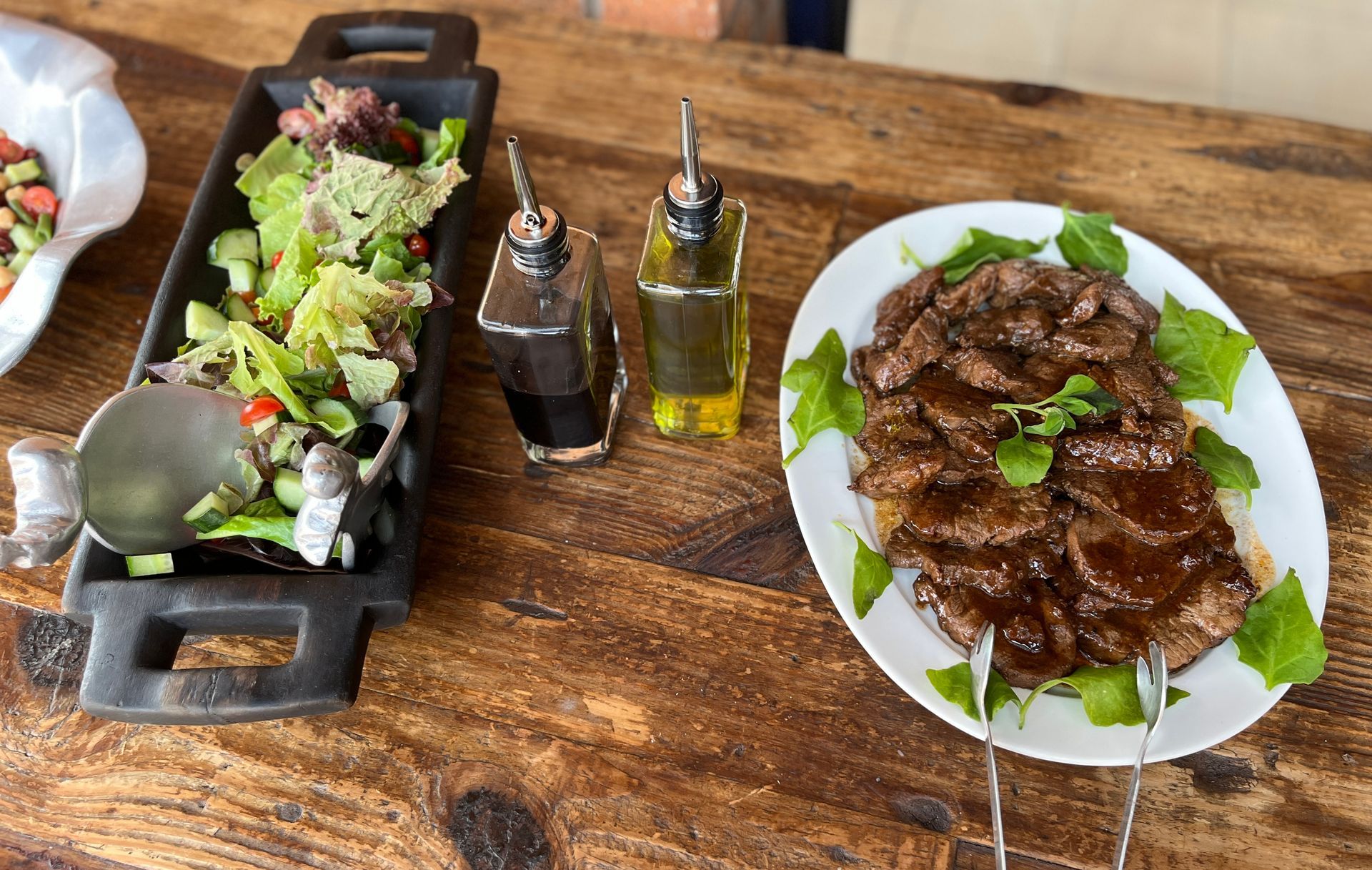 Plate of grilled meat on greens beside salad and olive oil on a wooden table