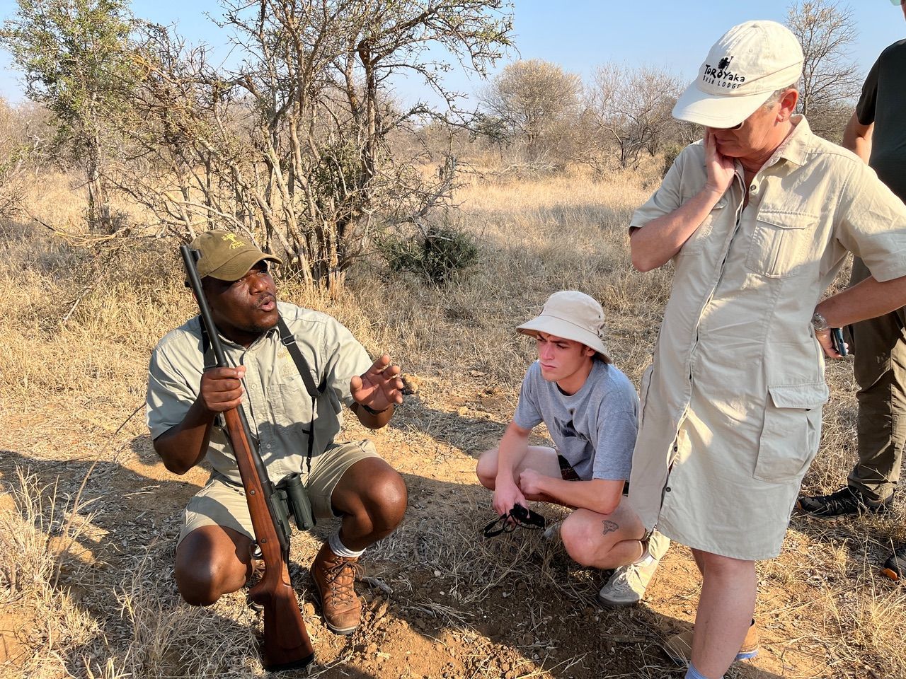 People gathered outdoors in dry brush, one man demonstrating a tool while two others watch nearby.