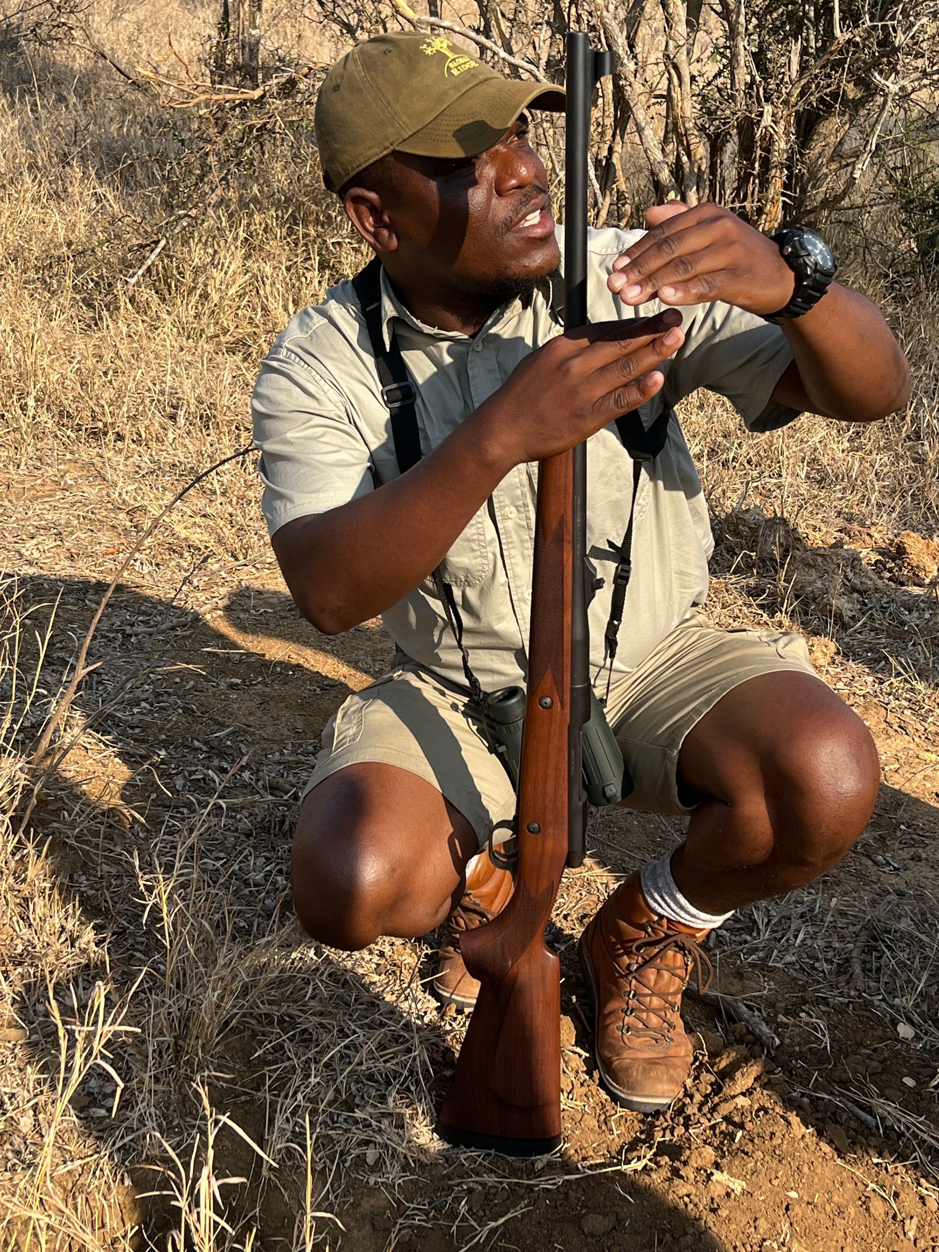 Man crouching outdoors, holding a rifle and adjusting the magazine in a dry grassy area.