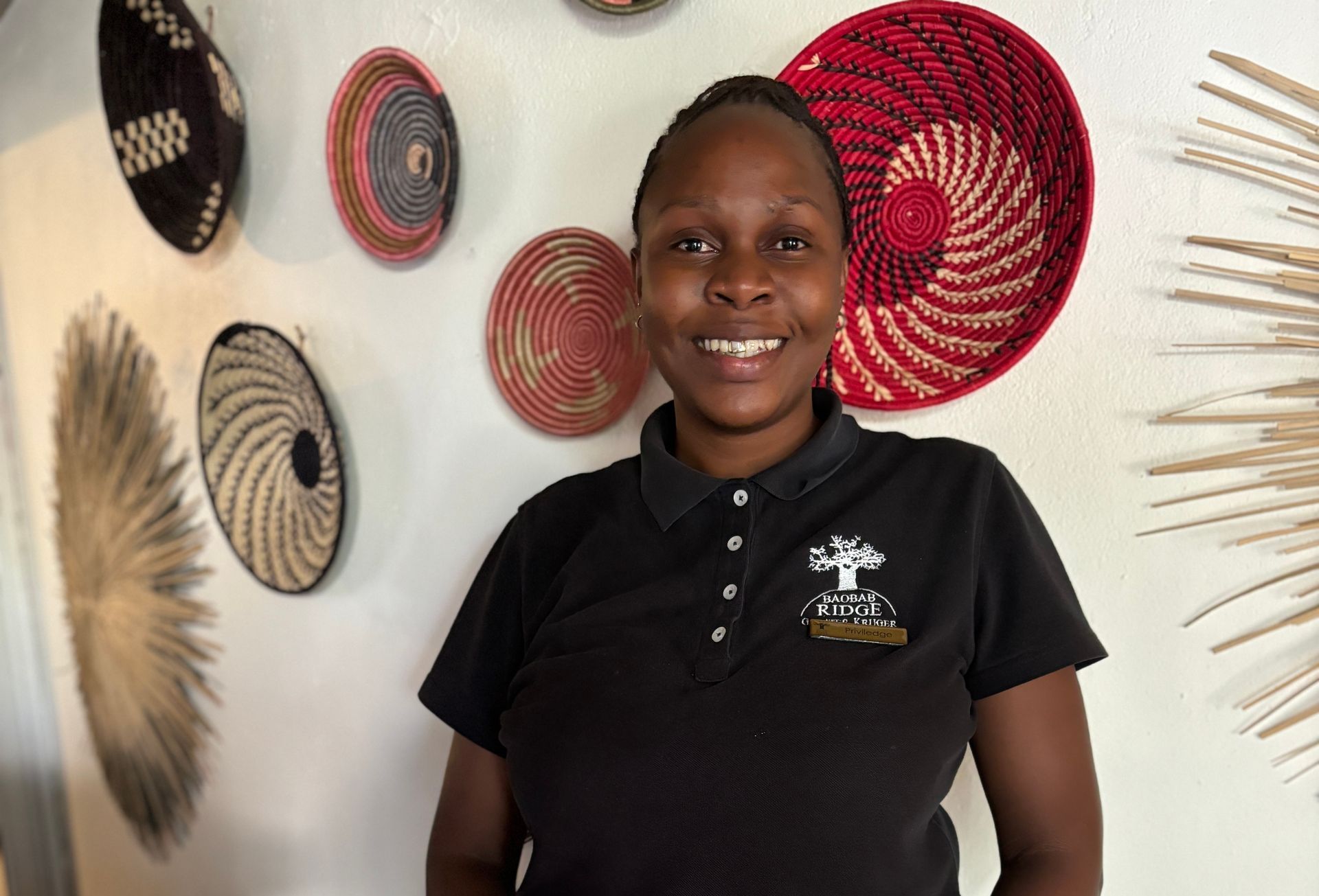 Person smiling indoors in a black polo, standing before a wall with colorful woven baskets and palm fronds
