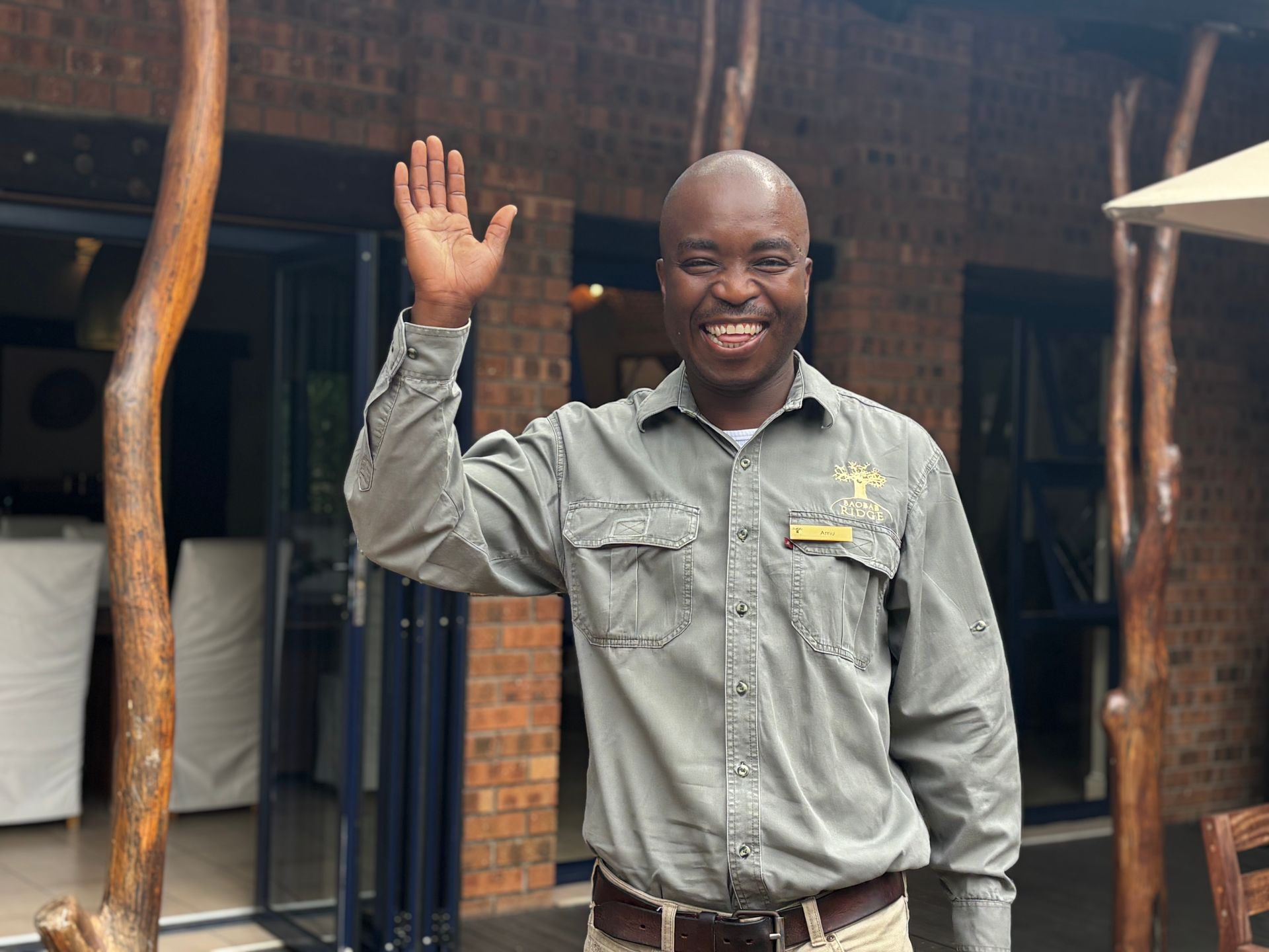 Smiling person in a gray shirt waving outside a brick building