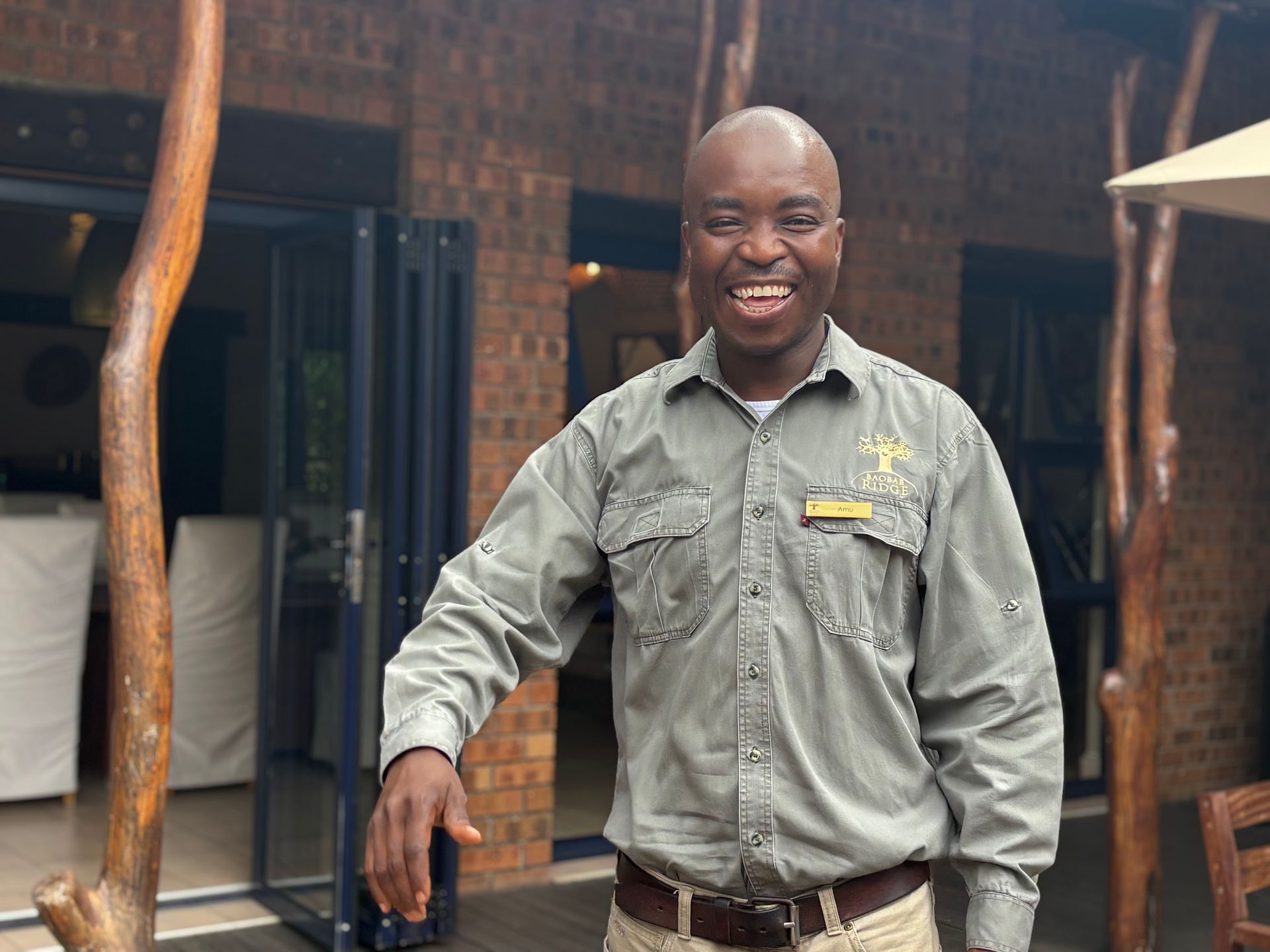 Smiling man in gray shirt standing outdoors beside a brick building and wooden posts