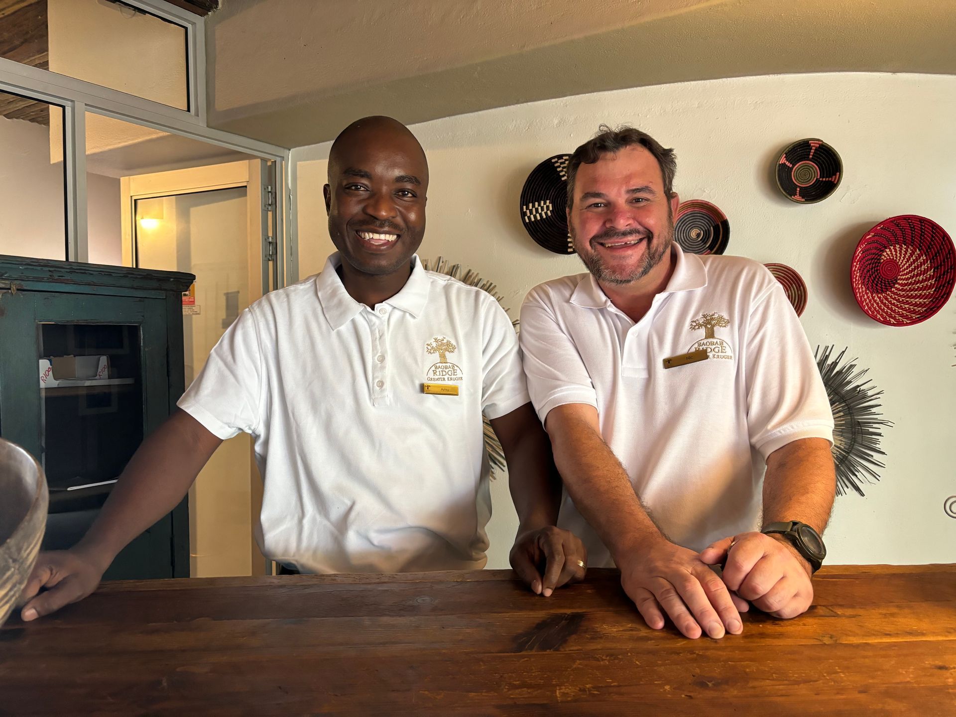 Two smiling men in white polos standing behind a wooden counter in a bright kitchen setting