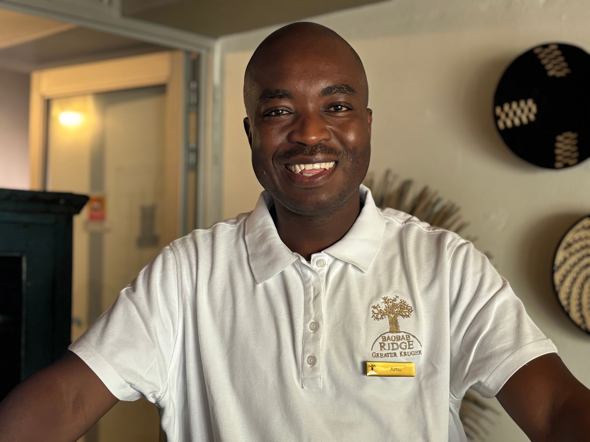 Smiling man in a white polo shirt standing indoors near a doorway, with wall decor in the background