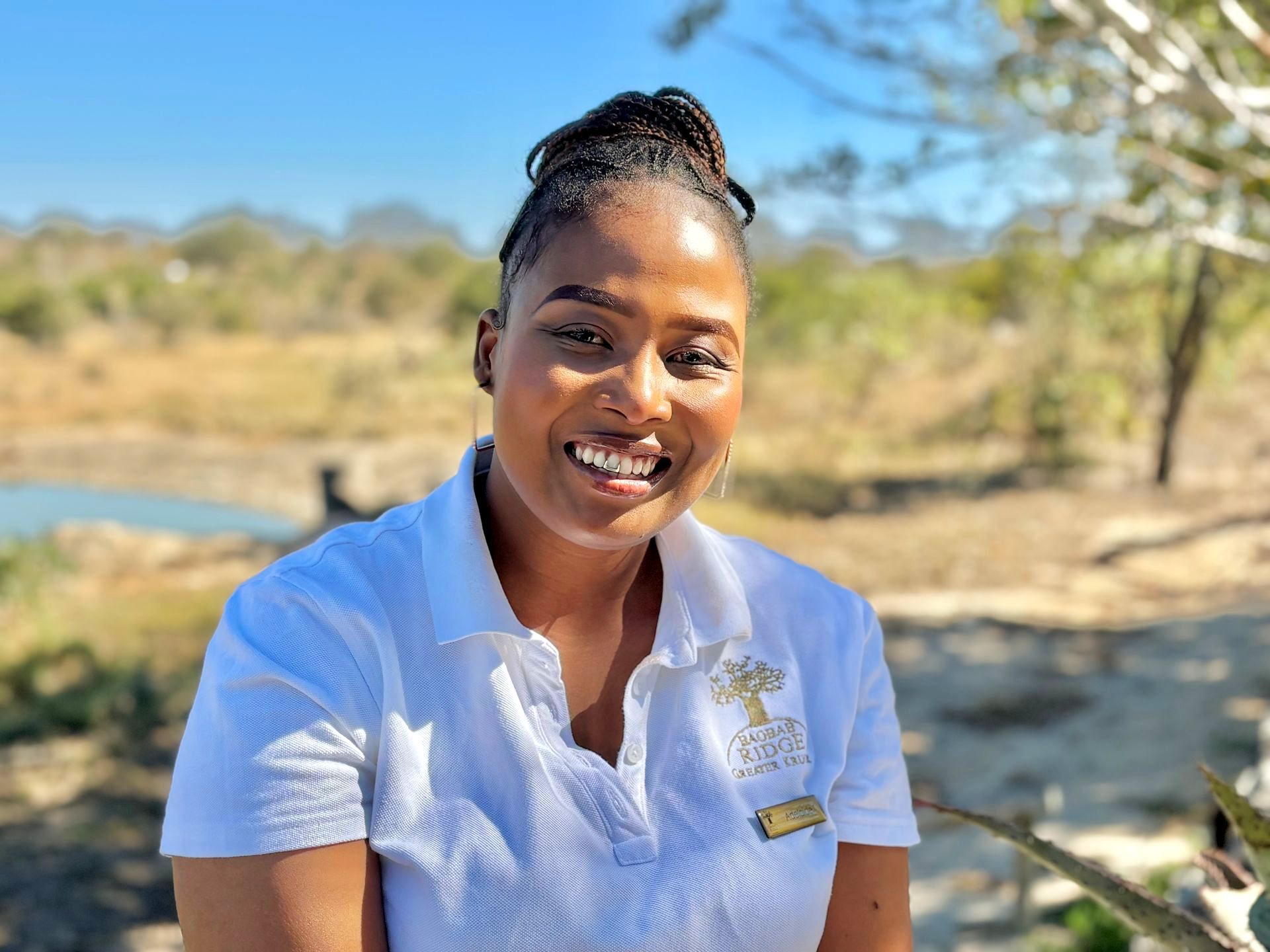 Smiling woman in a white polo shirt outdoors near a river under a bright blue sky