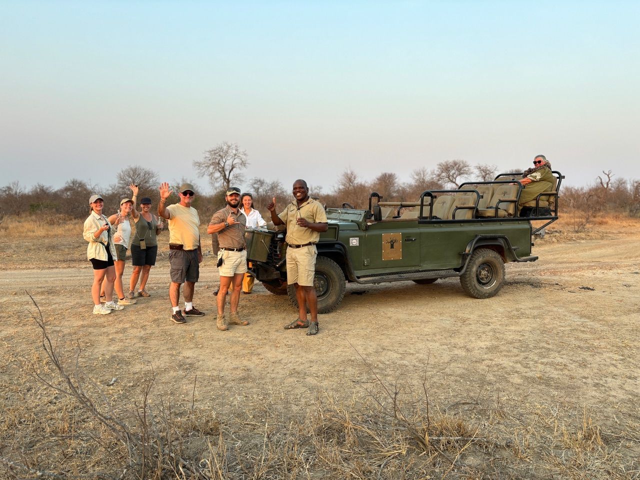 Group standing beside a green safari jeep in a dry field at sunset, with one person seated in the vehicle.