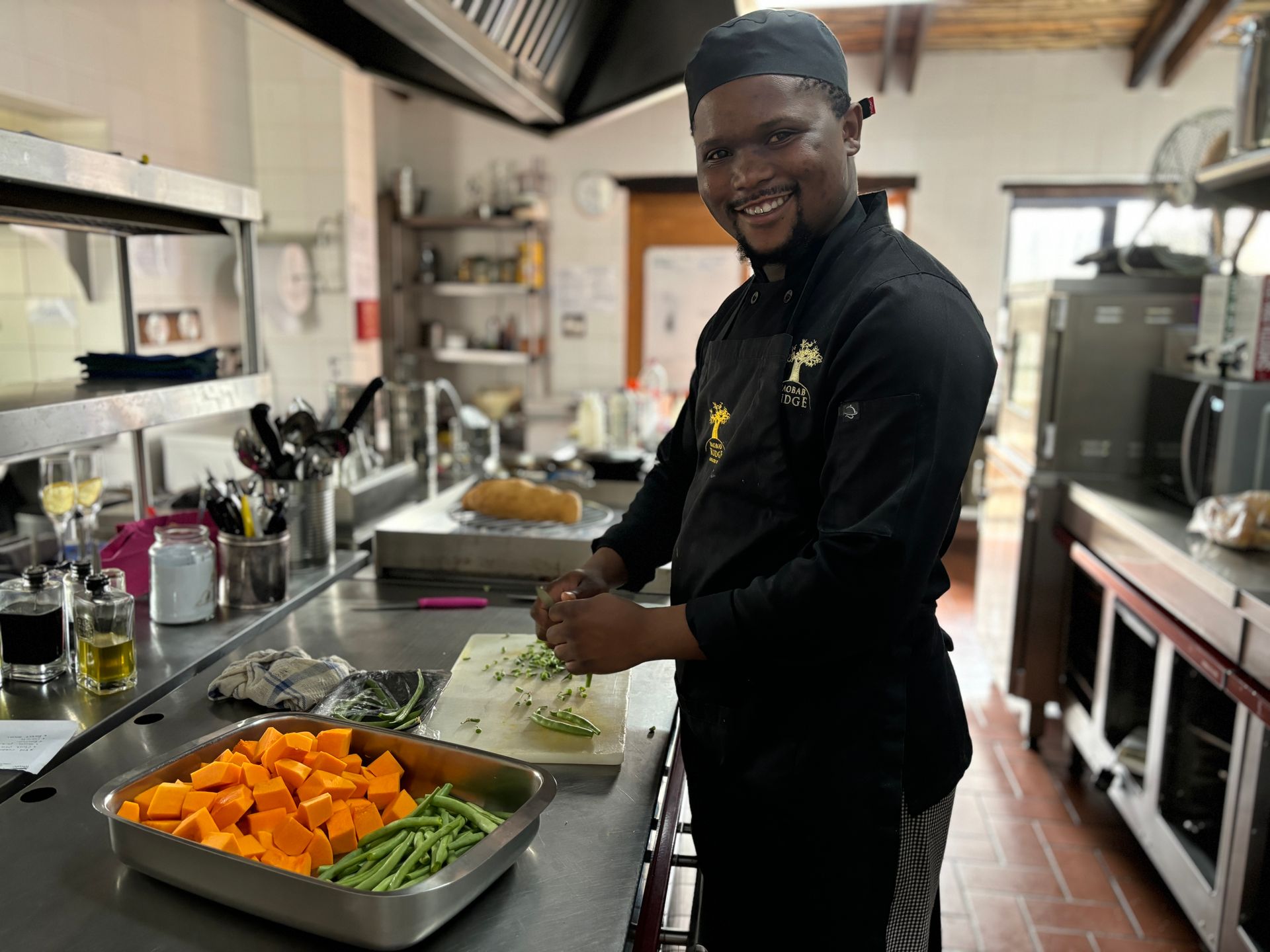 Chef smiling in a commercial kitchen, chopping vegetables beside trays of carrots and green beans.