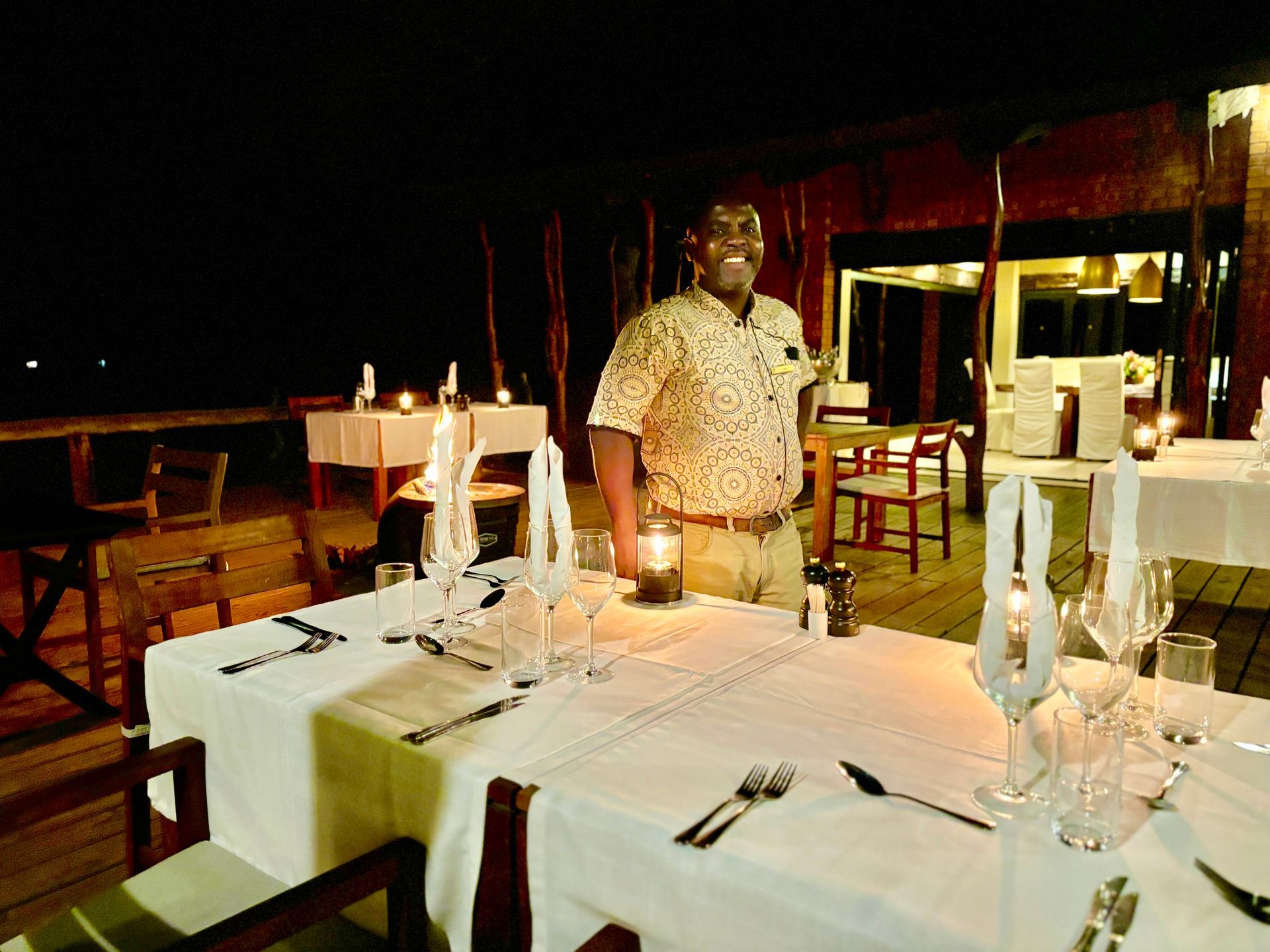 Candlelit restaurant table set for dinner, with a man standing beside it in a dimly lit dining room.