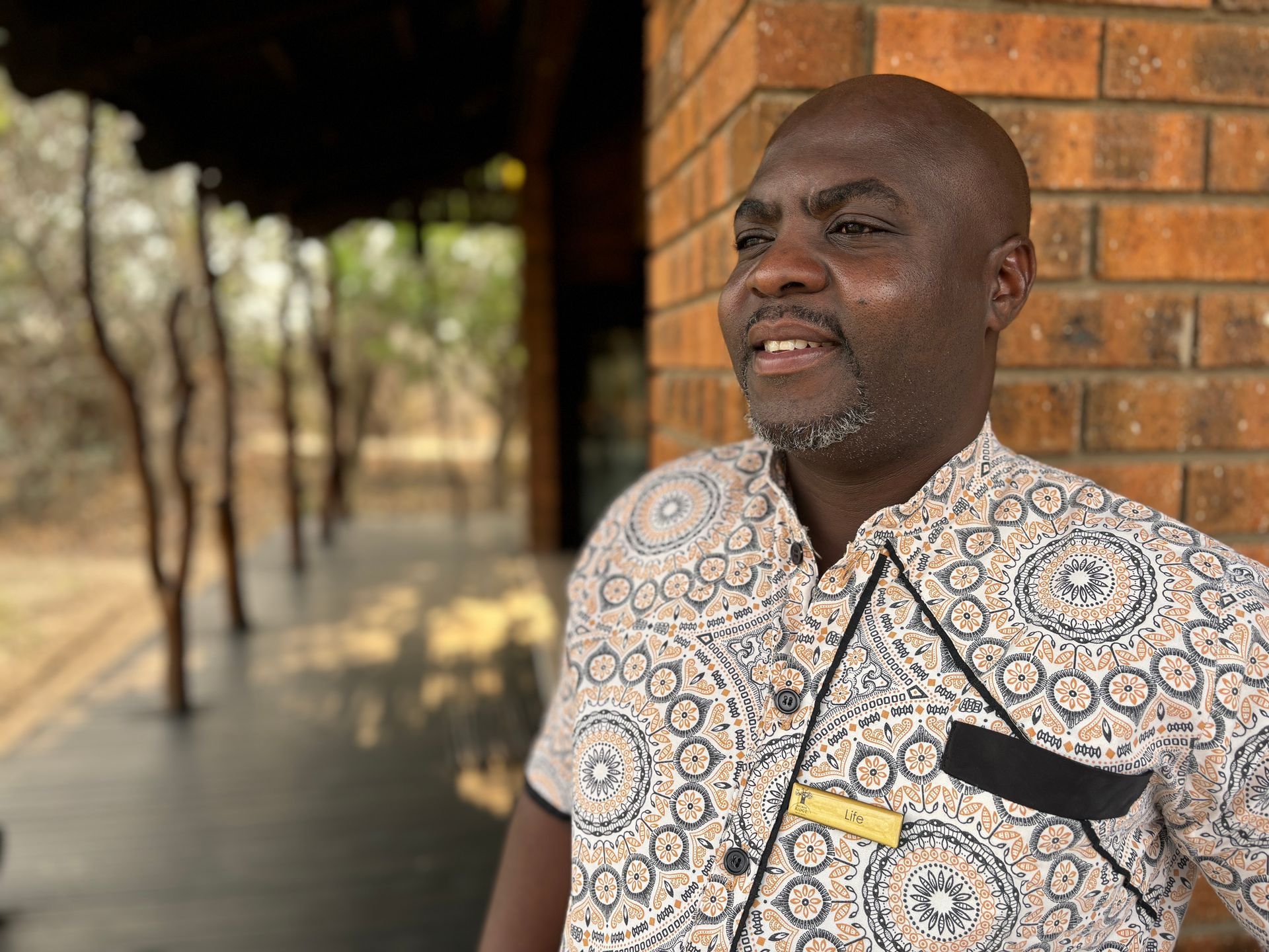 Man standing by a brick wall on a shaded walkway, wearing a patterned shirt and smiling.