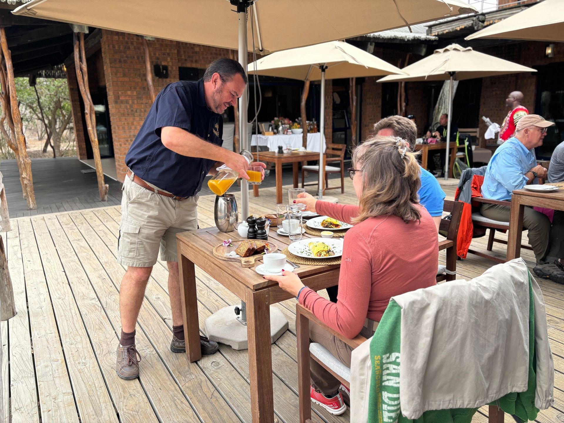 Man pouring orange drink at an outdoor café table with seated diners under umbrellas