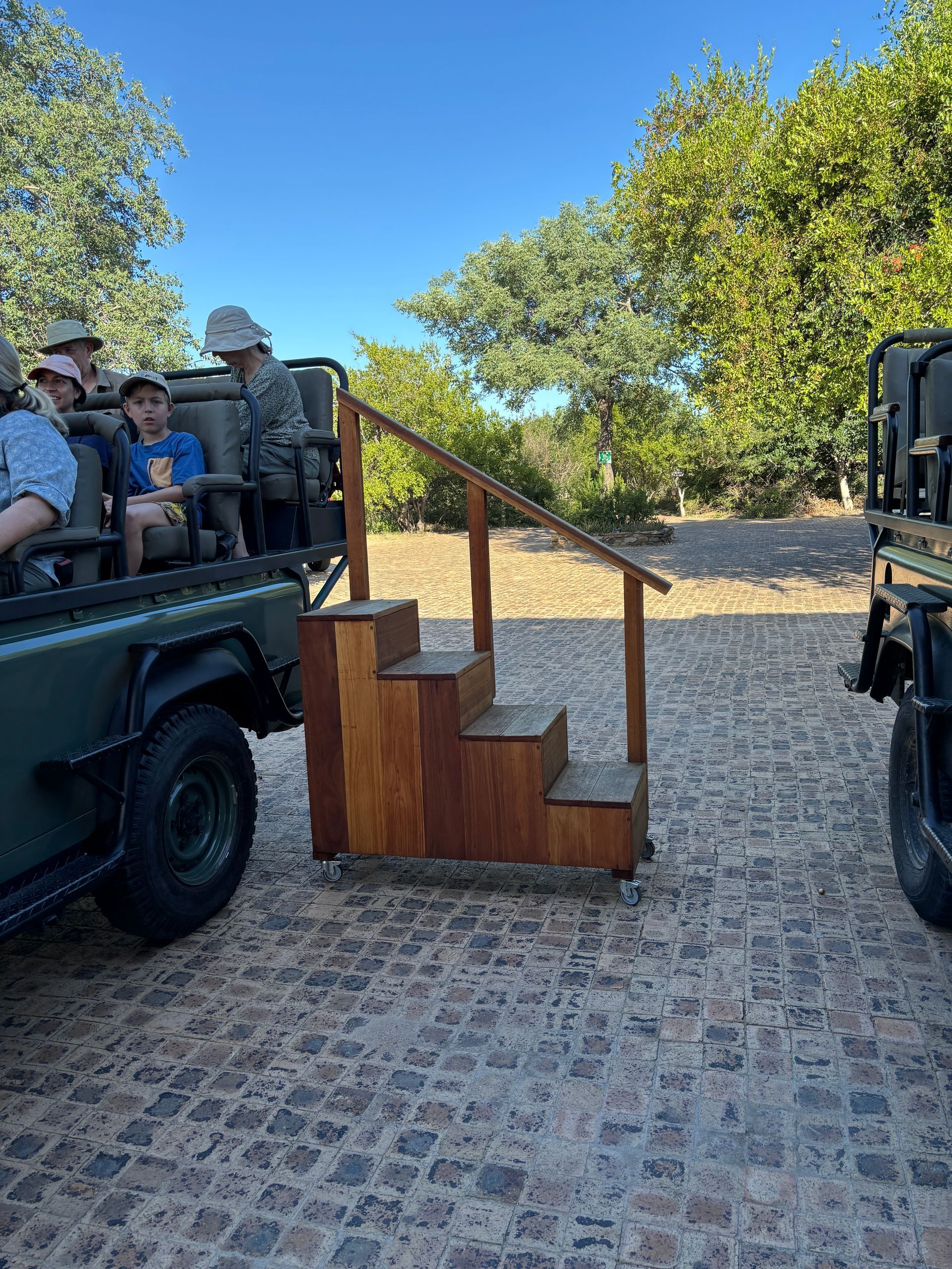 Open-sided safari vehicle with seated passengers beside a wooden step on a cobblestone road outdoors