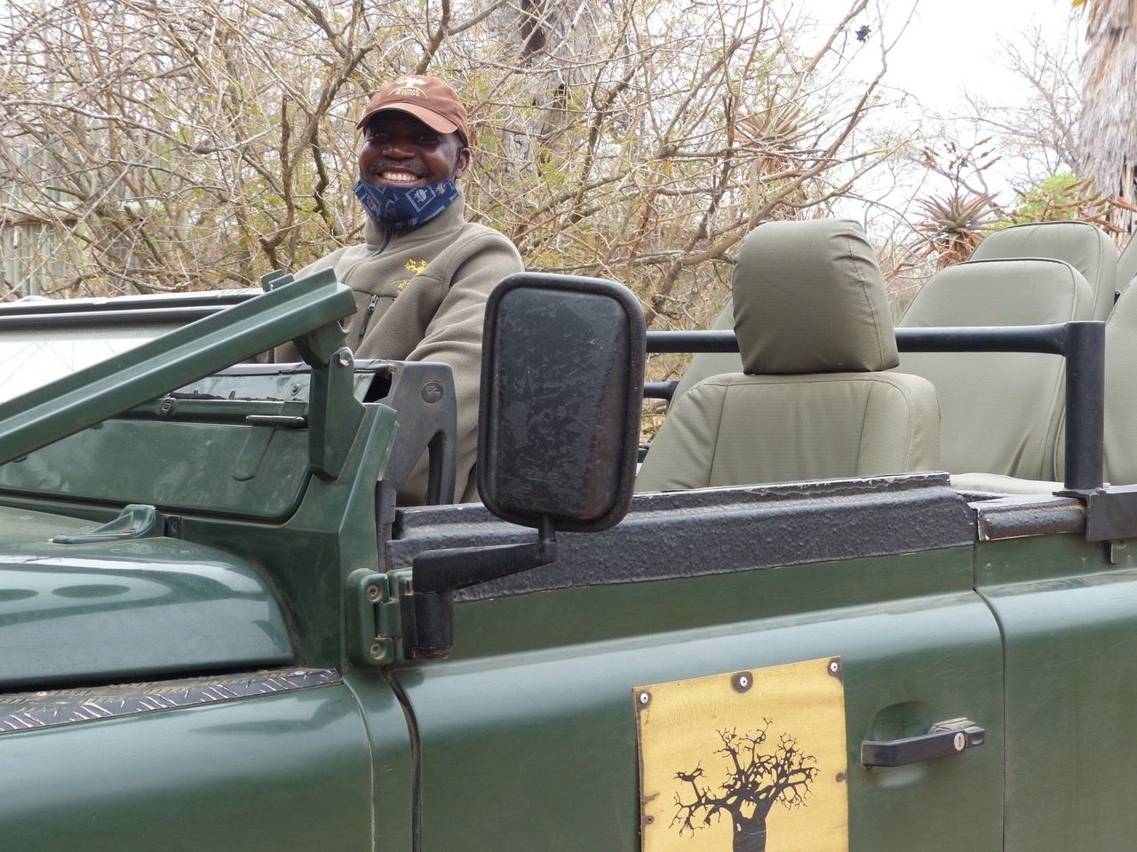 Person standing in green open-top vehicle, wearing a mask and cap, with bare trees in the background