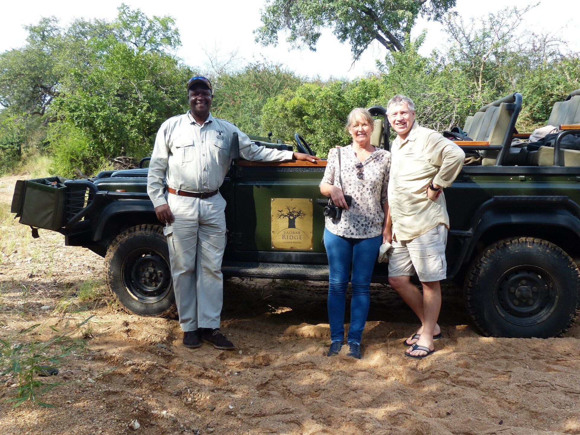 Three people standing beside a dark green safari vehicle in a muddy outdoor setting.