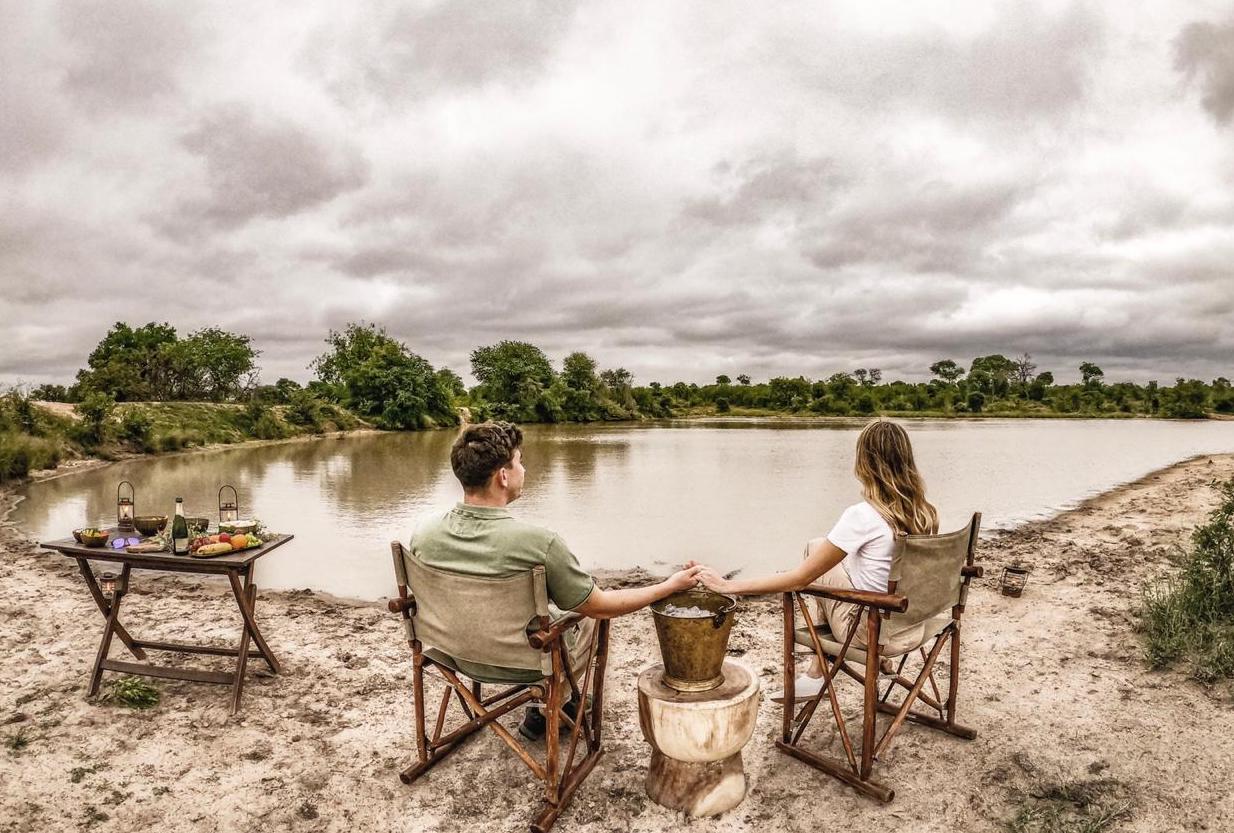 Two people relax in chairs by a muddy river, under a cloudy sky, with a small table beside them.