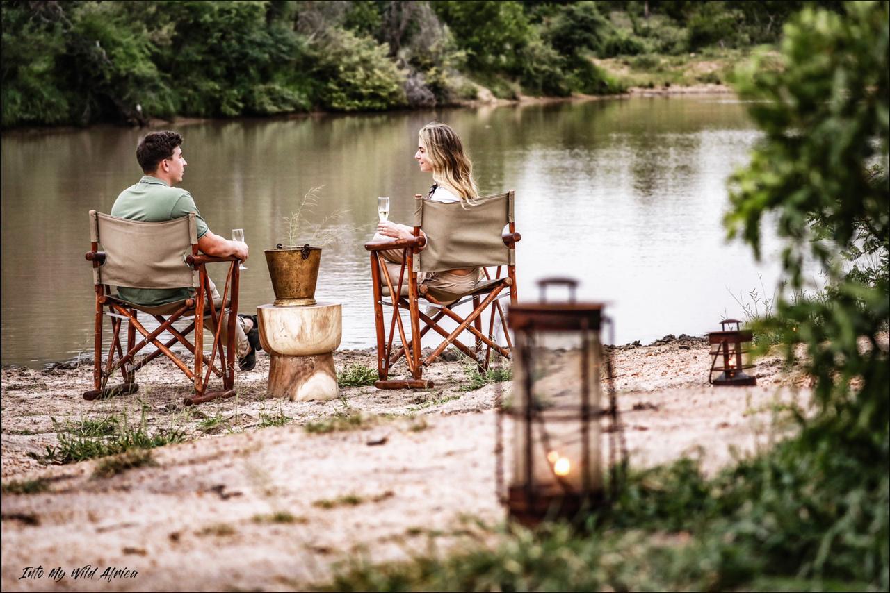 Two people sit in wooden chairs by a calm lakeside, with lanterns and a small table in the foreground.